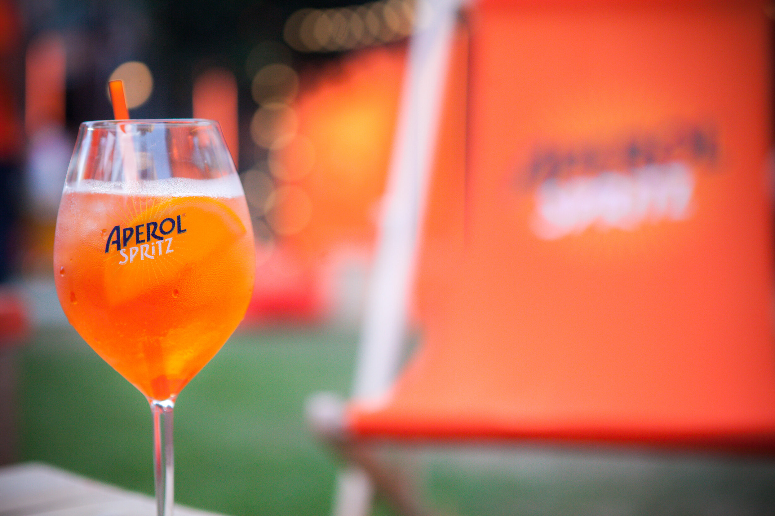 Verre d'Aperol Spritz avec une tranche d'orange et une paille orange, fond flou avec lumières et éléments oranges
