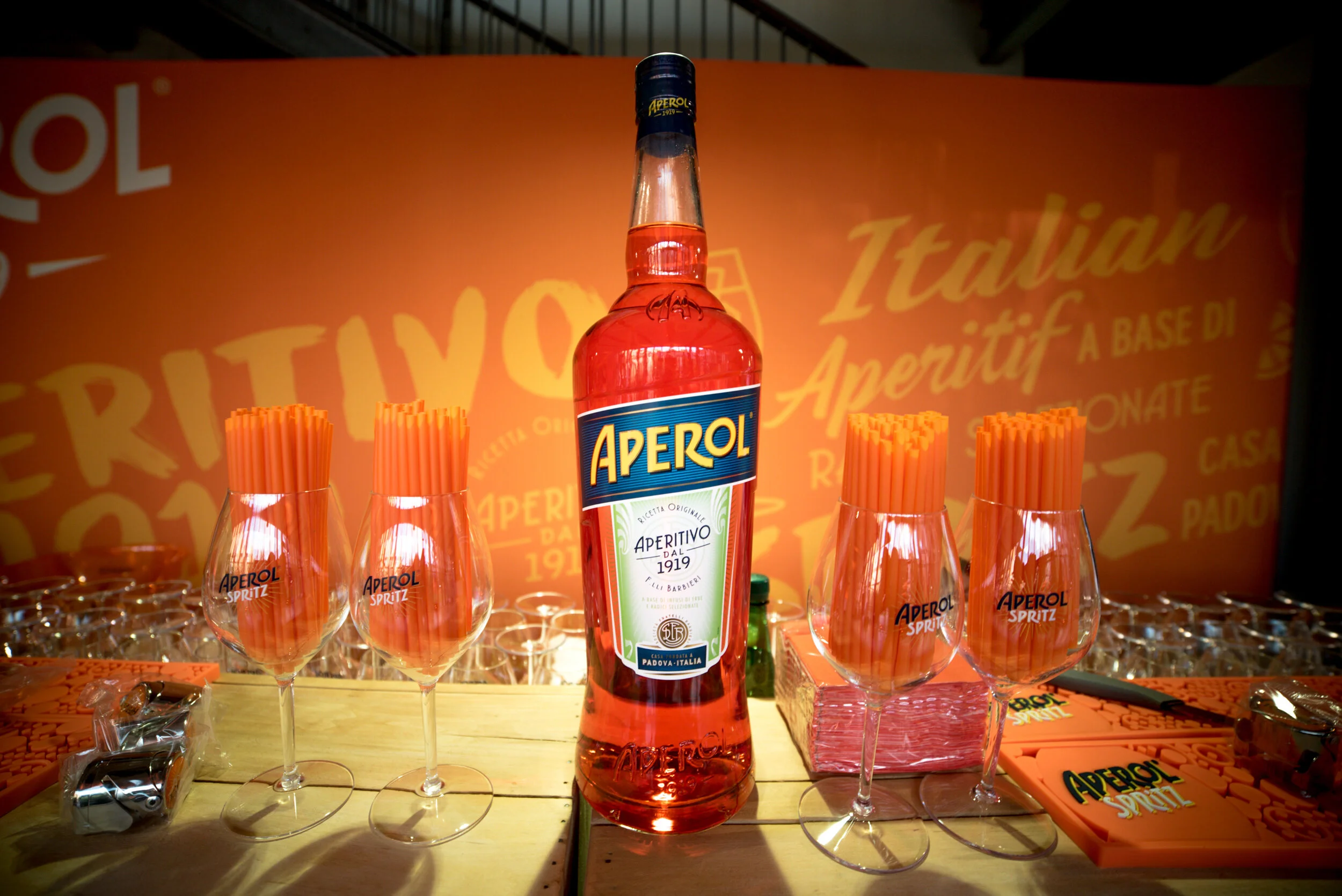 Une bouteille d'Aperol sur une table avec des verres contenant des pailles orange, ambiance de bar avec fond orange et logo Aperol.