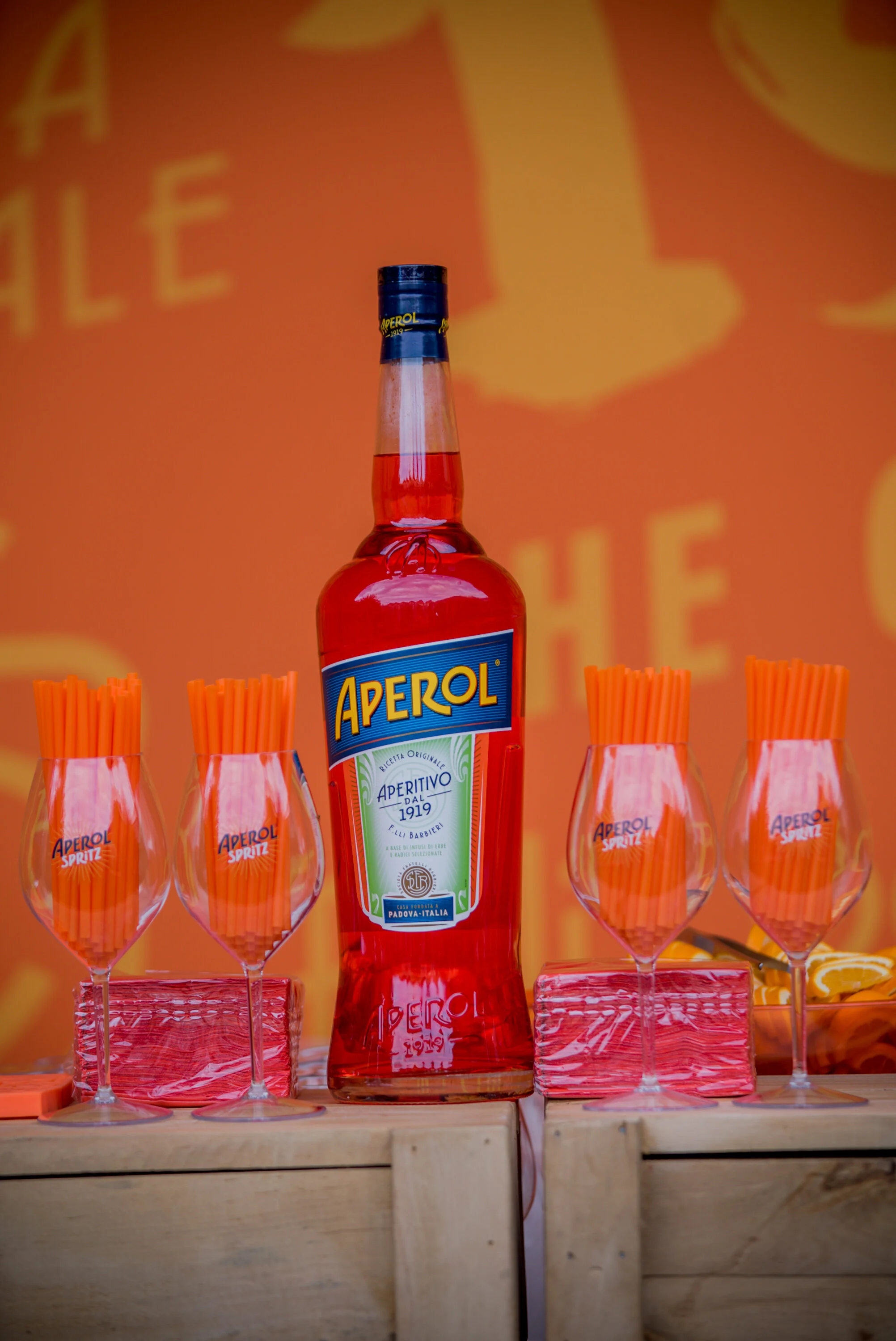Une bouteille d'Aperol entourée de quatre verres vides avec des pailles oranges, placés sur une surface en bois, avec un fond orange.