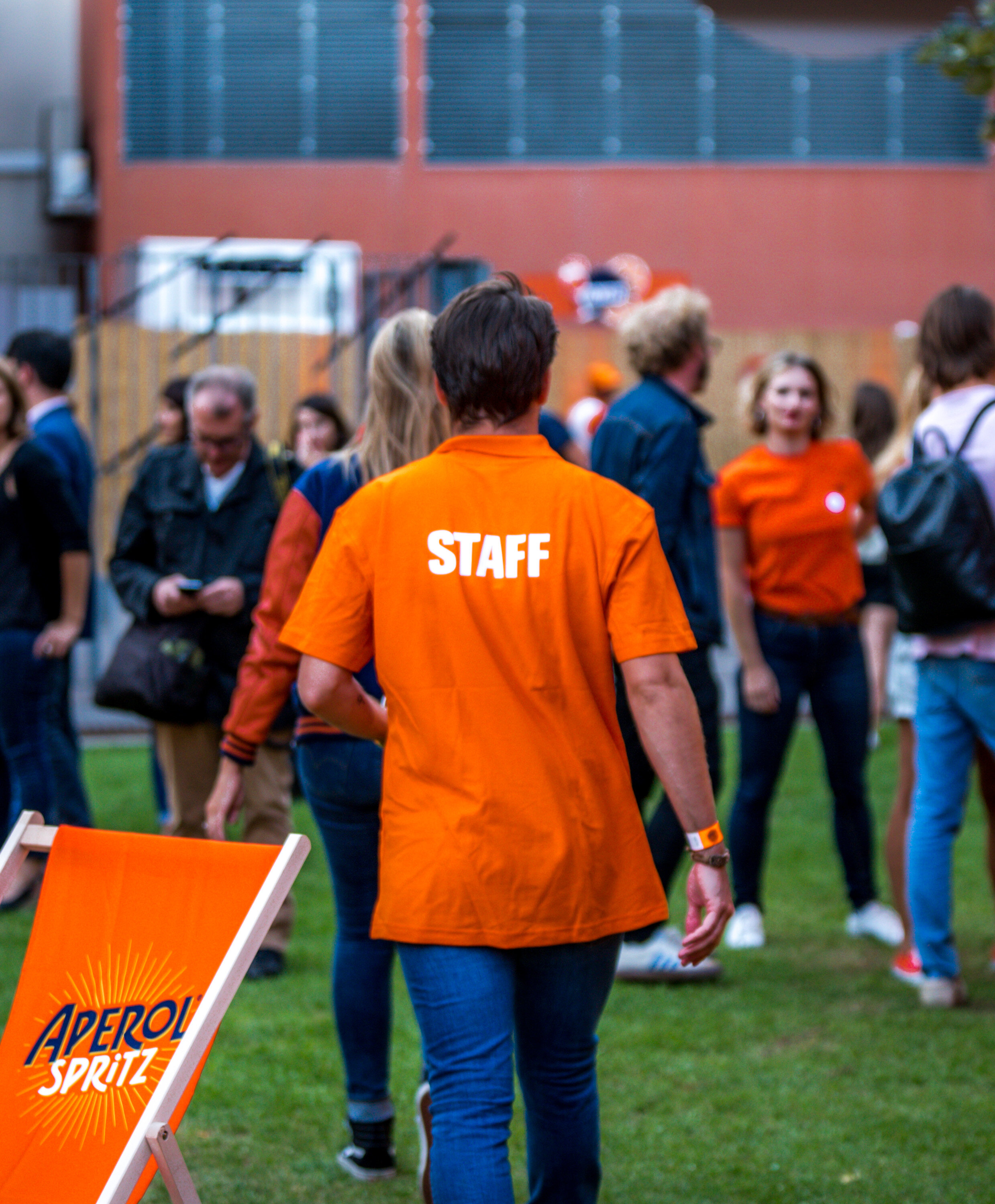 Une personne portant un t-shirt orange avec l'inscription 'STAFF' vue de dos, entourée de plusieurs autres personnes dont certaines portent des t-shirts orange, dans un environnement extérieur en soirée.