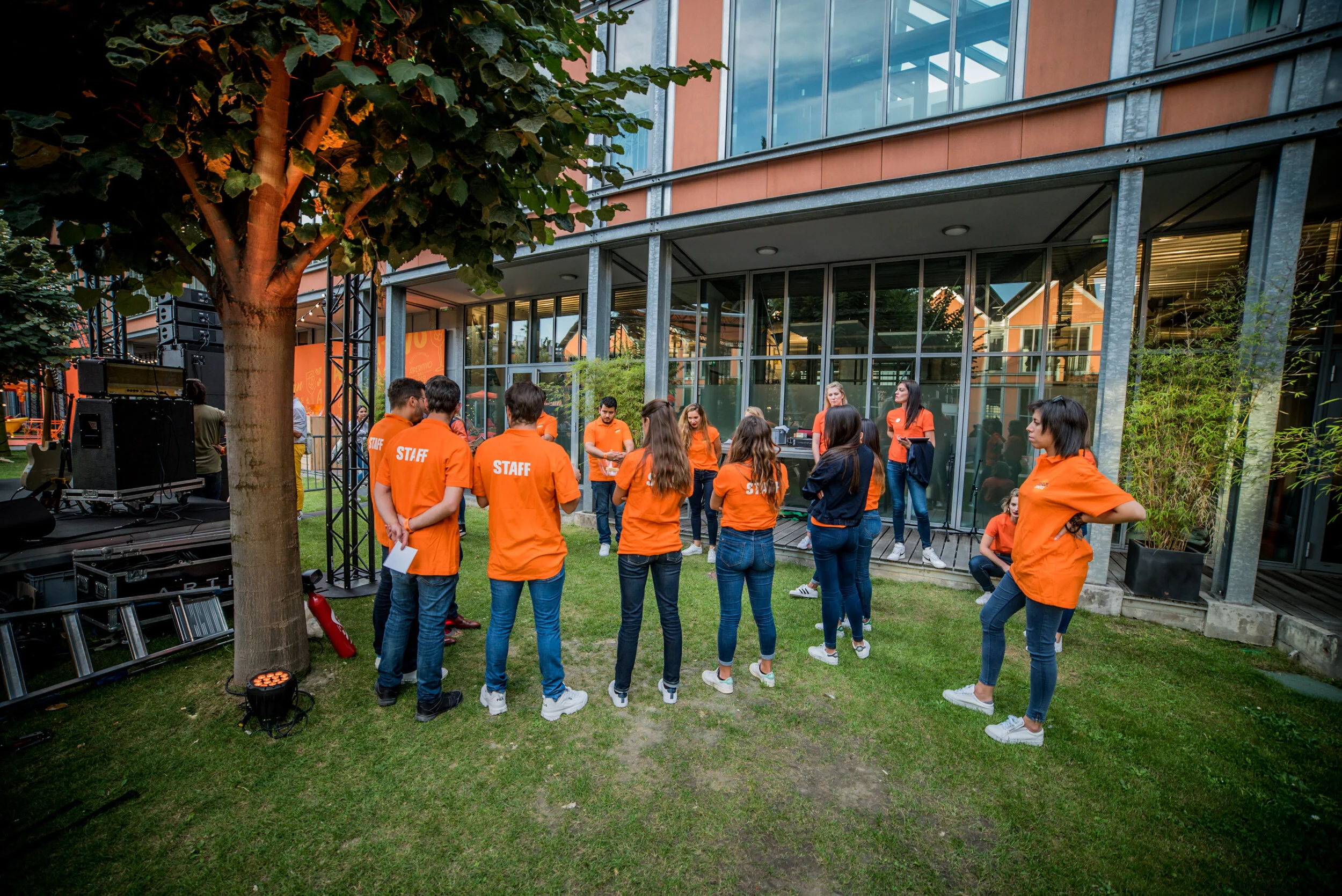Groupe de personnes en tenue orange avec le mot 'STAFF' lors d'une activité extérieure près d'un bâtiment moderne avec beaucoup de fenêtres.