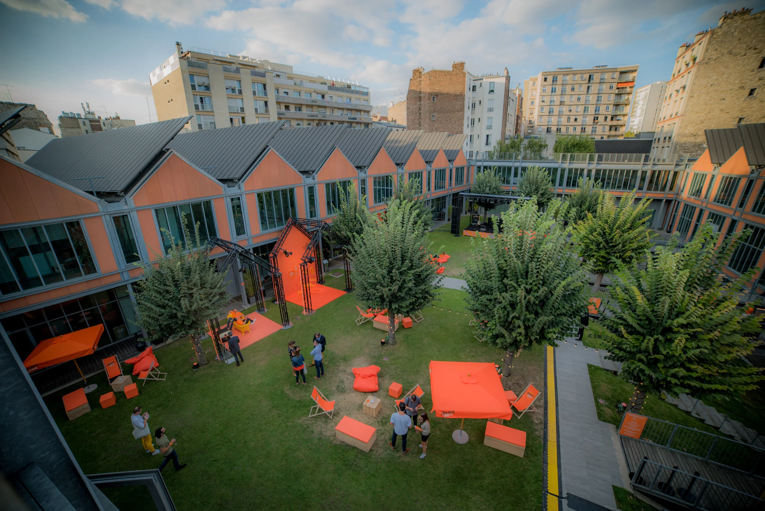 Cour intérieure d'un immeuble moderne avec arbres, canapés orange, parasols et personnes discutant dans un espace vert.