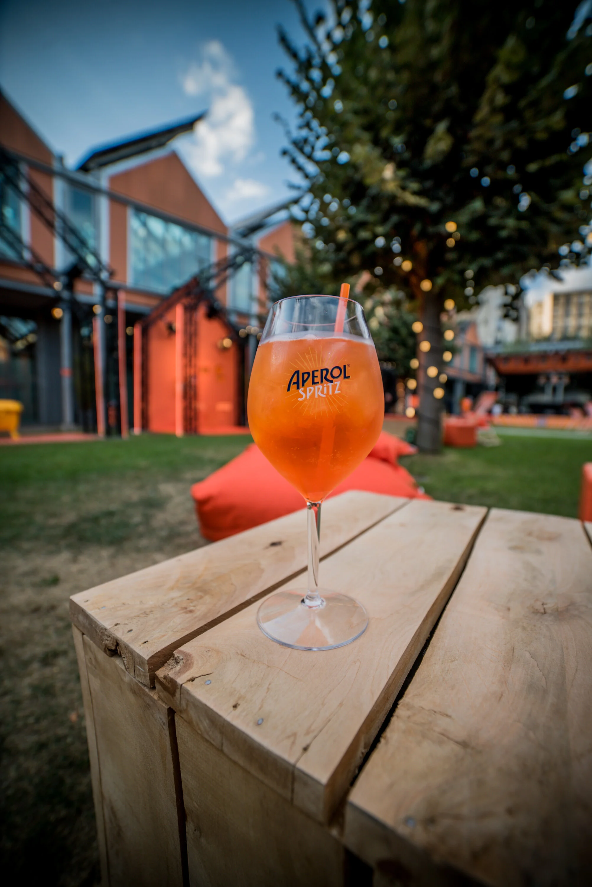 Verre à cocktail Aperol Spritz sur une table en bois, extérieur avec maison colorée et arbres décorés de lumières