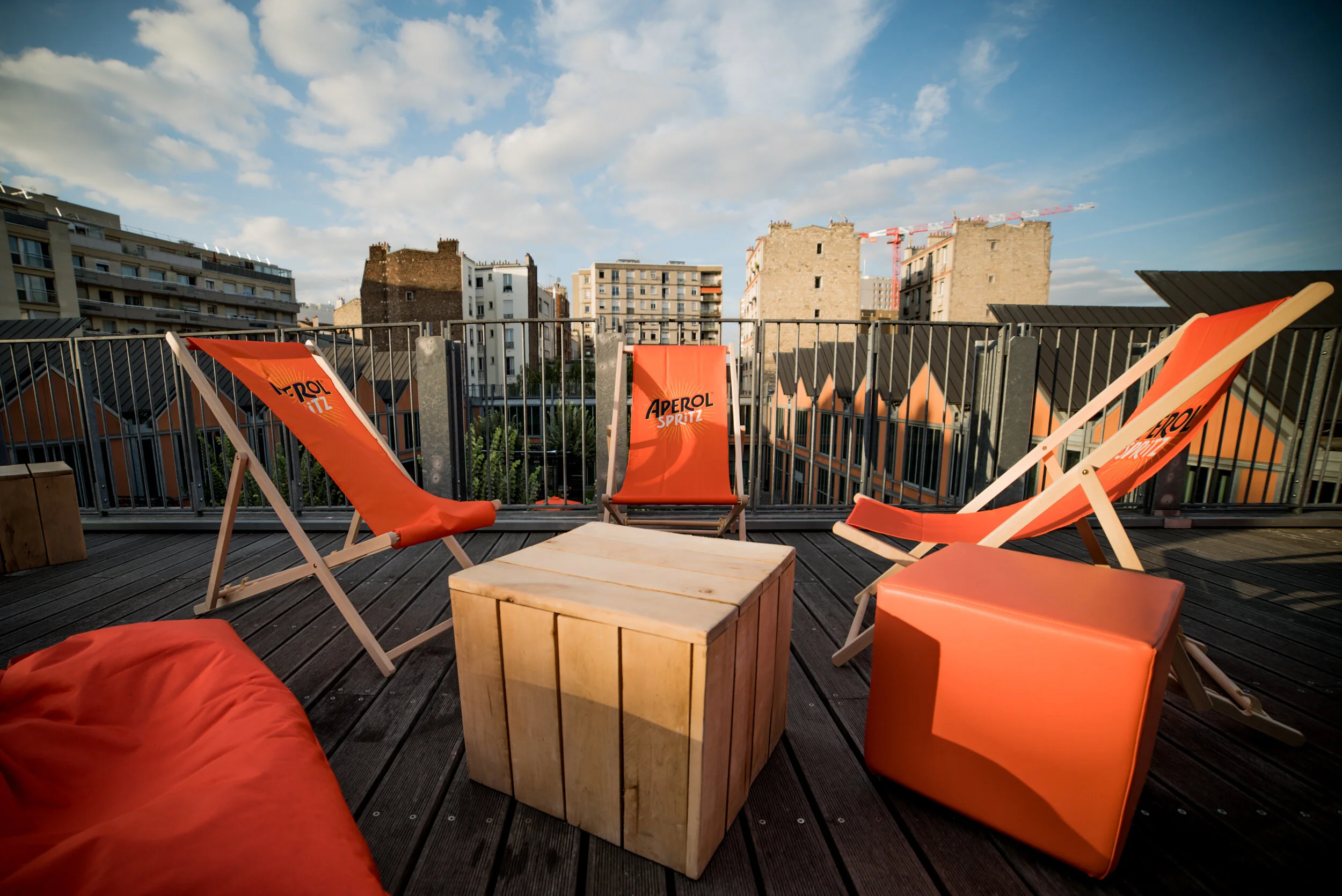 Terrasse avec chaises longues oranges, un cube en bois, un coussin rouge, et une vue urbaine avec des bâtiments et un ciel partiellement nuageux.