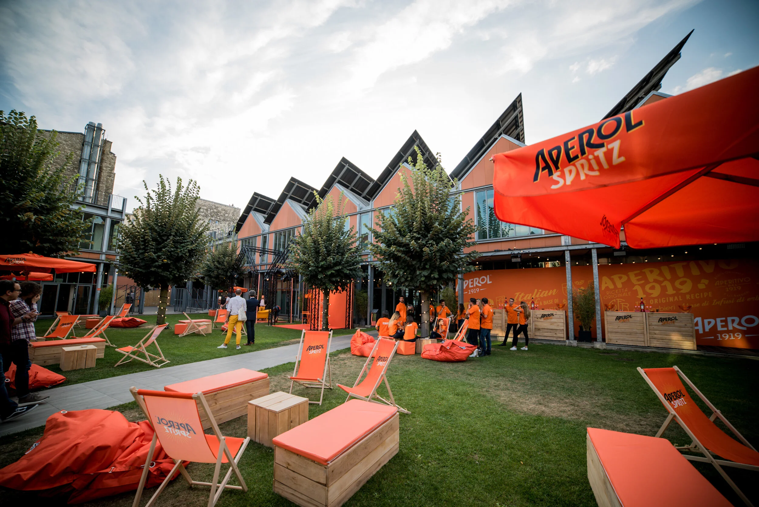 Balconnières modernes en maison, espaces de détente avec chaises longues orange et coussins, arbres arborant guirlandes lumineuses, un événement extérieur avec accueil et personnel en orange, décoration de branding Aperol Spritz, ambiance estivale en