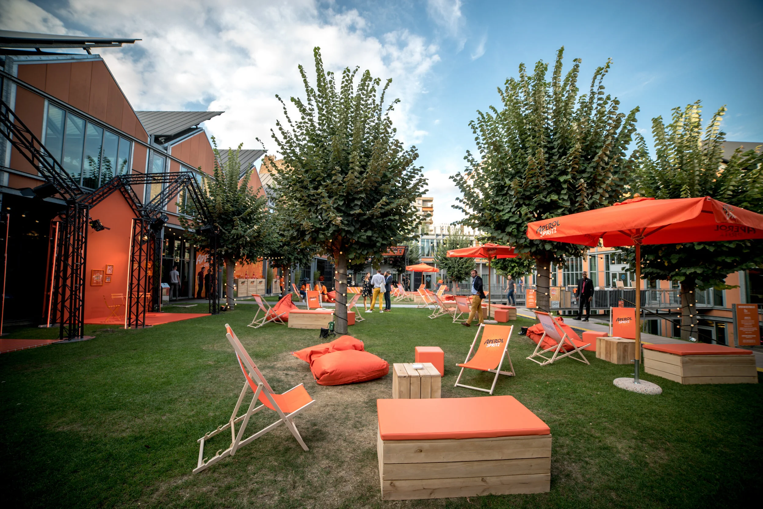 Espace extérieur avec des chaises longues orange, parasols orange, arbres verts, herbe verte, structures en métal noir, bâtiments modernes, personnes se promenant, ambiance de jour ensoleillé.