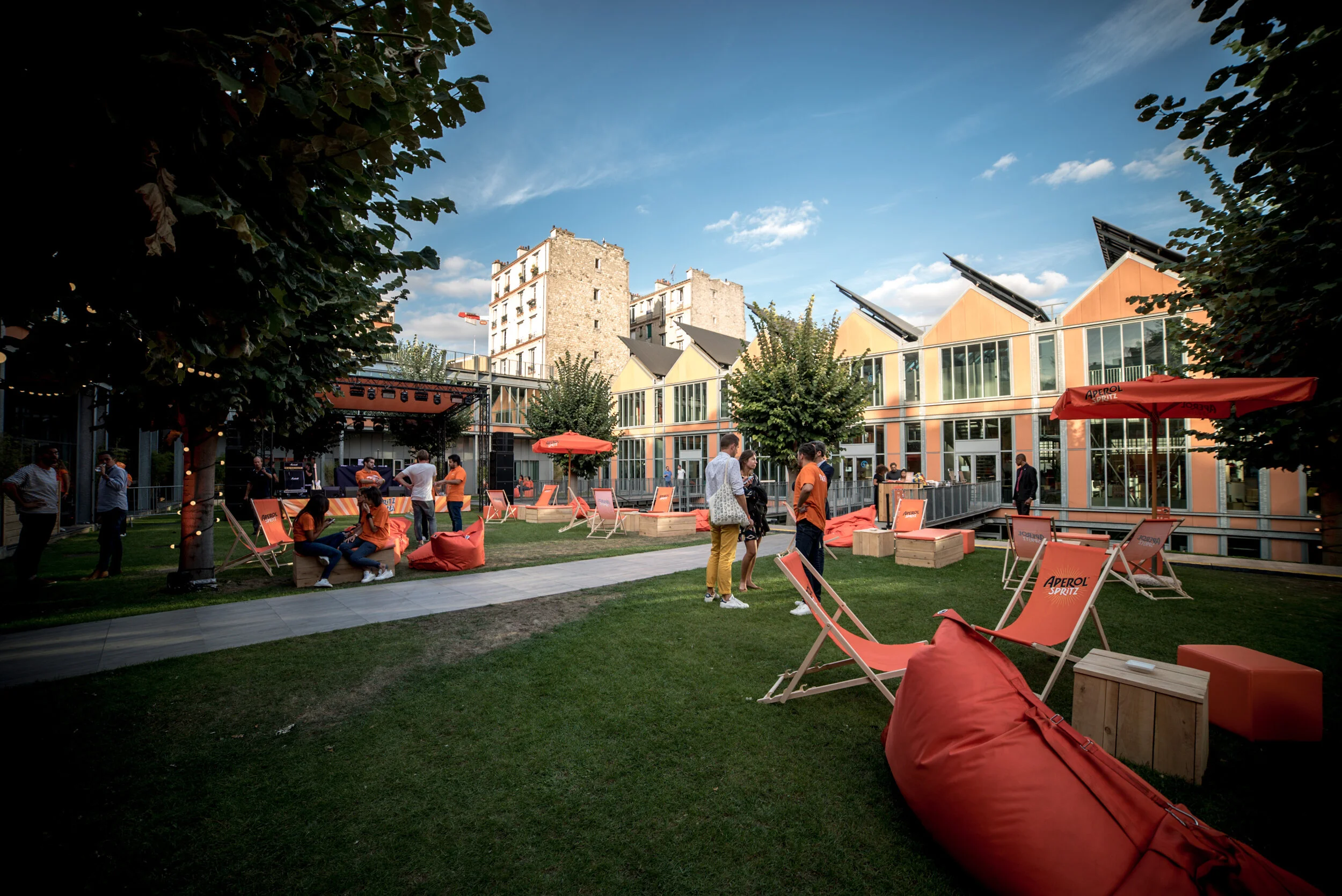 Une cour extérieure animée avec des gens discutant, des chaises longues orange et des parasols, dans un bâtiment moderne avec beaucoup de fenêtres, sous un ciel bleu en fin d'après-midi.