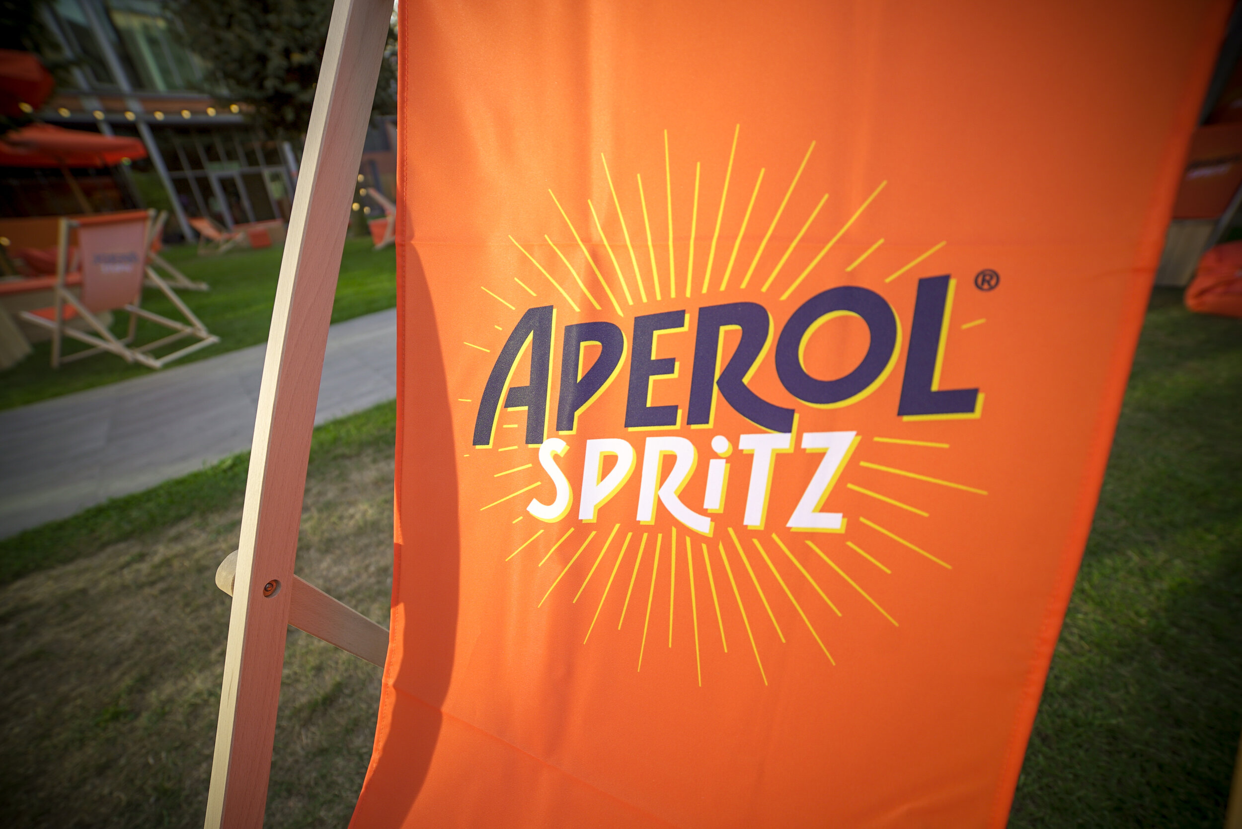 Une bannière orange avec le logo 'AEROL SPRITZ' en caractères bleus et blancs, entourée de rayons jaunes, dans un espace extérieur avec des chaises longues en toile orange et un chemin en pierre au fond.