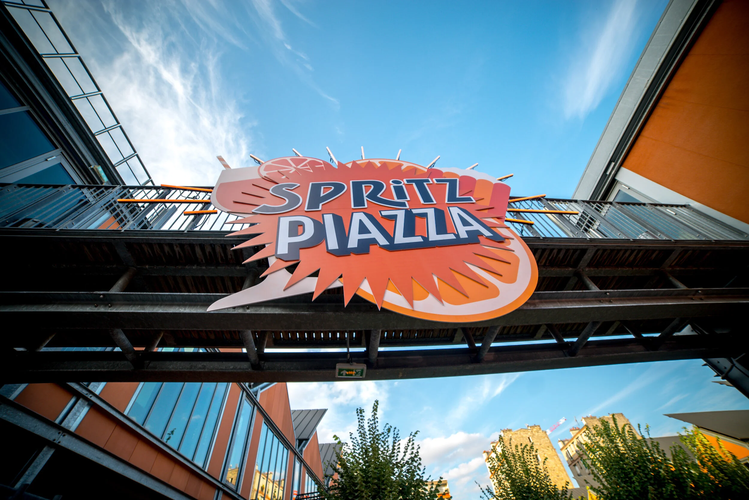 Panneau lumineux avec le texte "Spritz Piazza" suspendu entre deux bâtiments, avec un ciel bleu et des arbres en arrière-plan.