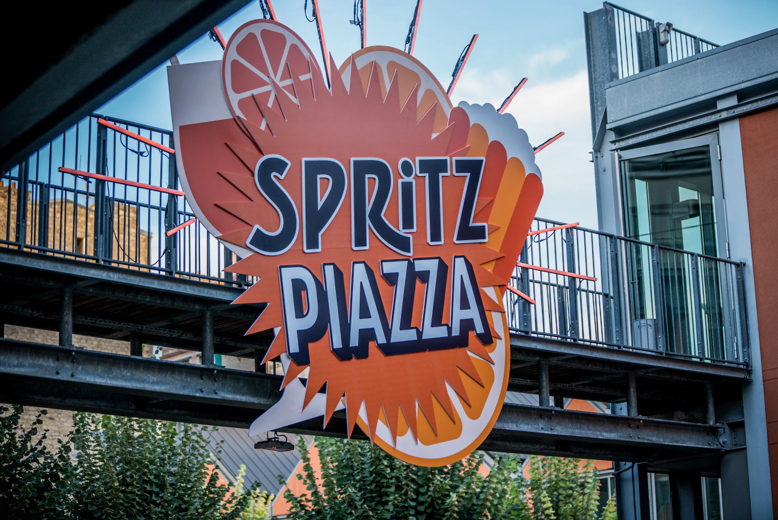 Un grand panneau en forme de symbole d'une tranche d'orange avec le texte 'Spritz Piazza' écrit dessus, suspendu au-dessus d'un espace public en plein air.