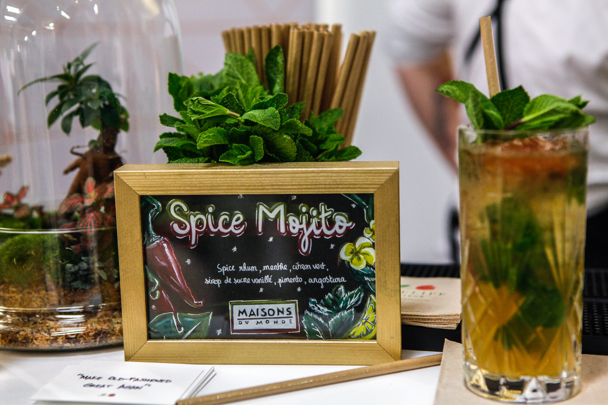 Un tableau noir avec écrit 'Spice Mojito', entouré d'illustrations de piments, de feuilles de menthe, et de citrons, placé devant des plantes, avec un mojito dans un verre à côté.