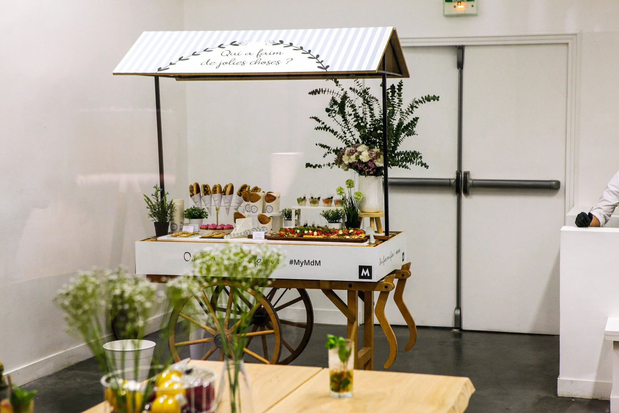 Stand de vente de gourmandises dans une foire, avec un toit en forme de petite maison. Le stand est décoré avec des plantes et des fleurs, et propose divers desserts, notamment des éclairs, dans un cadre intérieur lumineux.