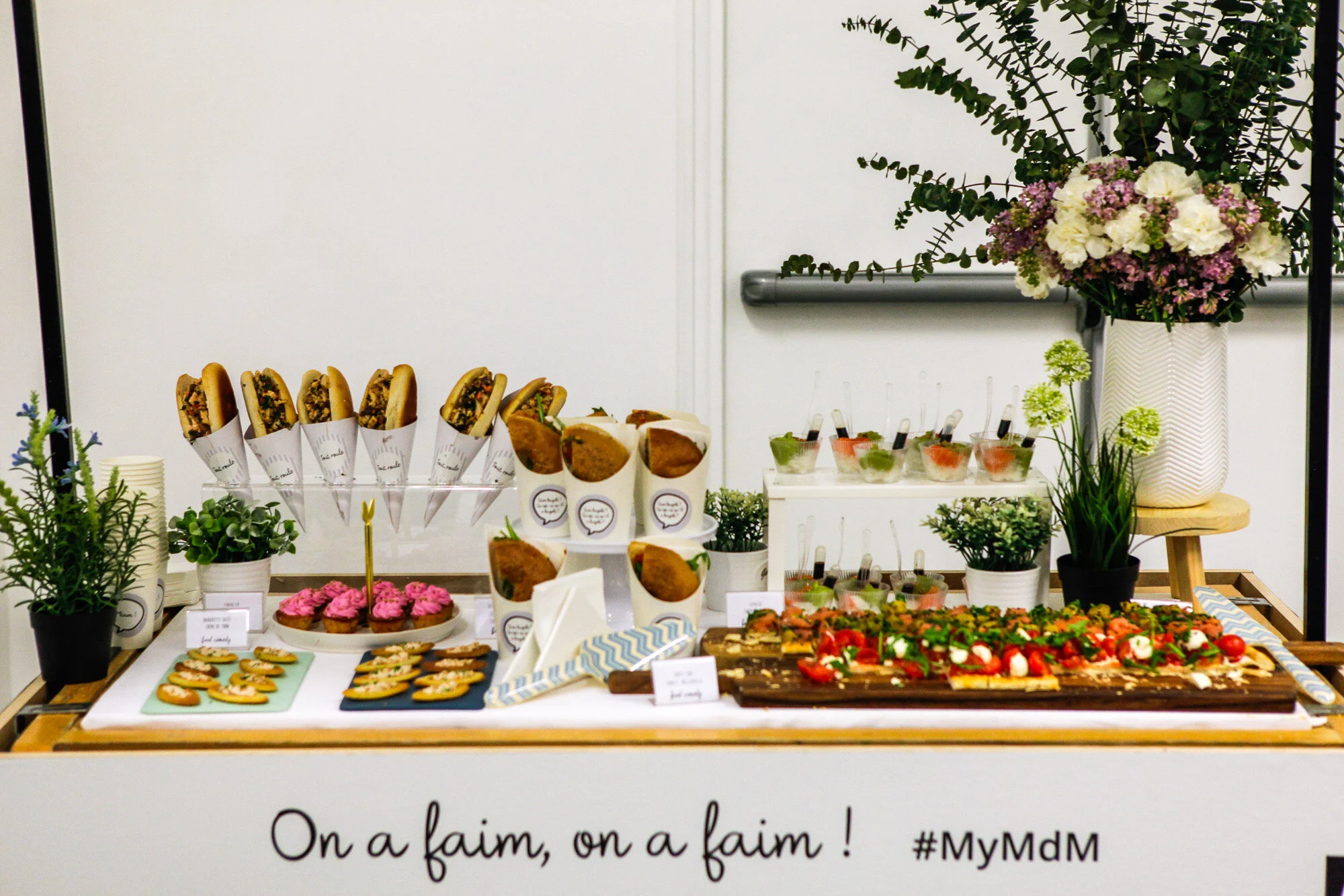 Buffet avec sandwich, cupcakes, cookies, et antipasti, décoré avec des plantes et des fleurs dans un pot blanc, inscription en français "On a faim, on a faim !".