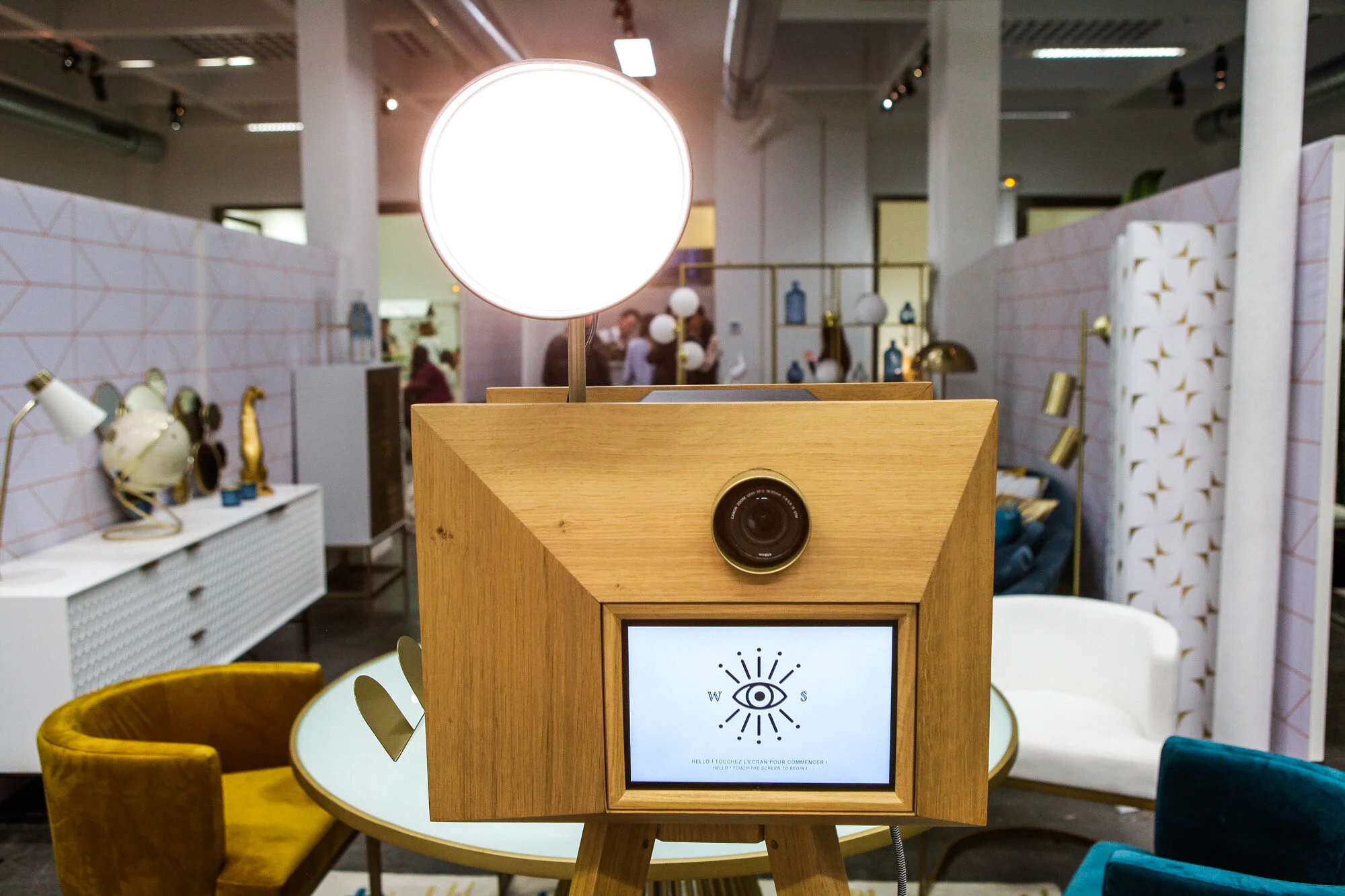 Photobooth en bois avec un écran et une caméra, éclairé par une lampe ronde blanche en haut, dans un espace intérieur décoré avec des meubles et des lampes design.