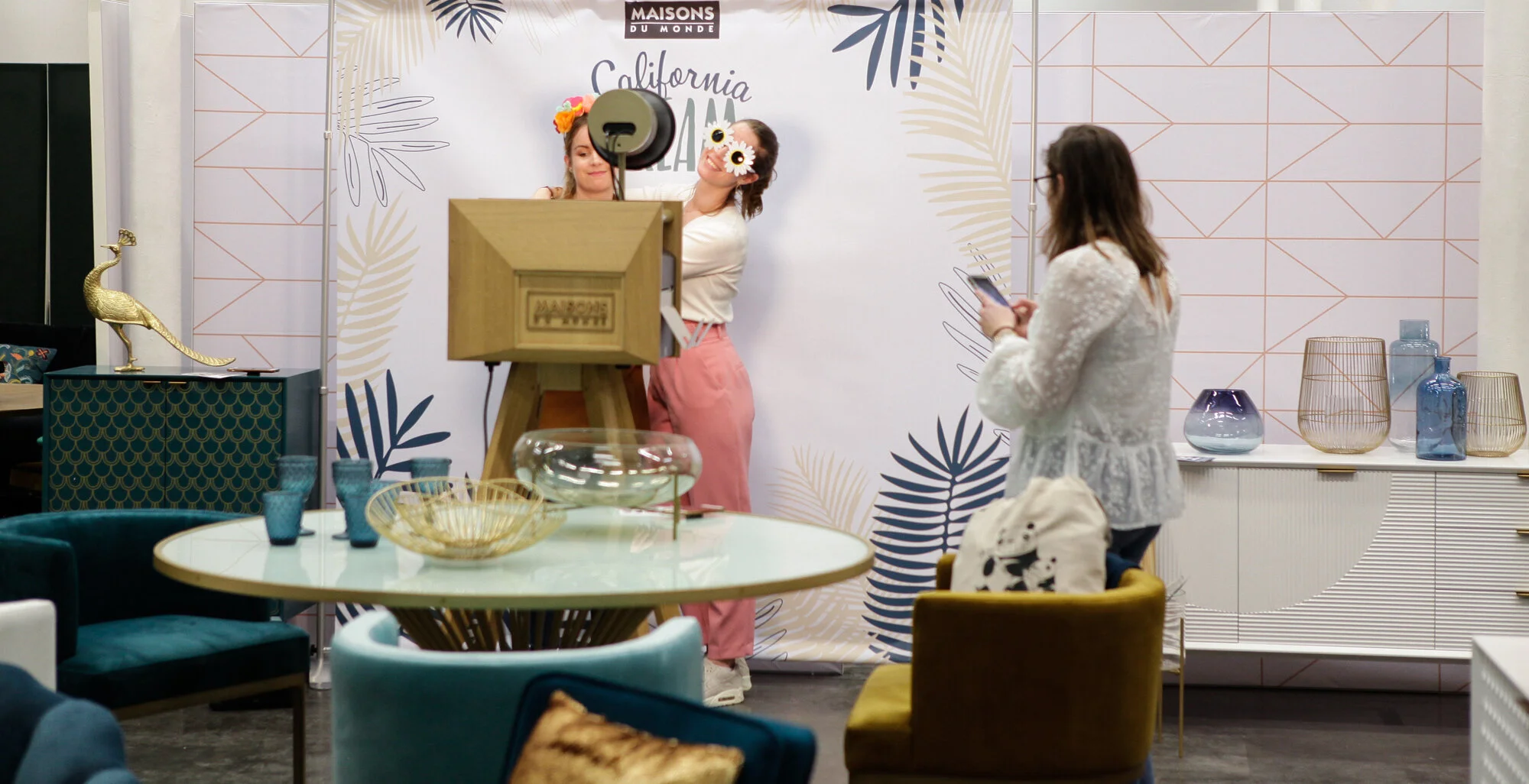 Deux femmes posant pour une photo dans un décor coloré avec des objets de décoration et une table avec des verres, une fille en arrière-plan porte des accessoires amusants et une autre femme regarde son téléphone.