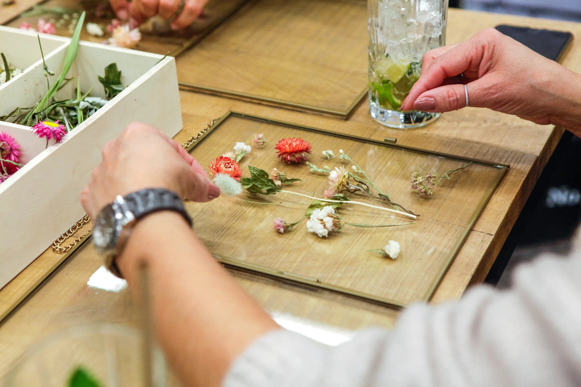 Une personne assemble des fleurs séchées sur une surface en bois, avec une boîte de fleurs séchées à proximité.