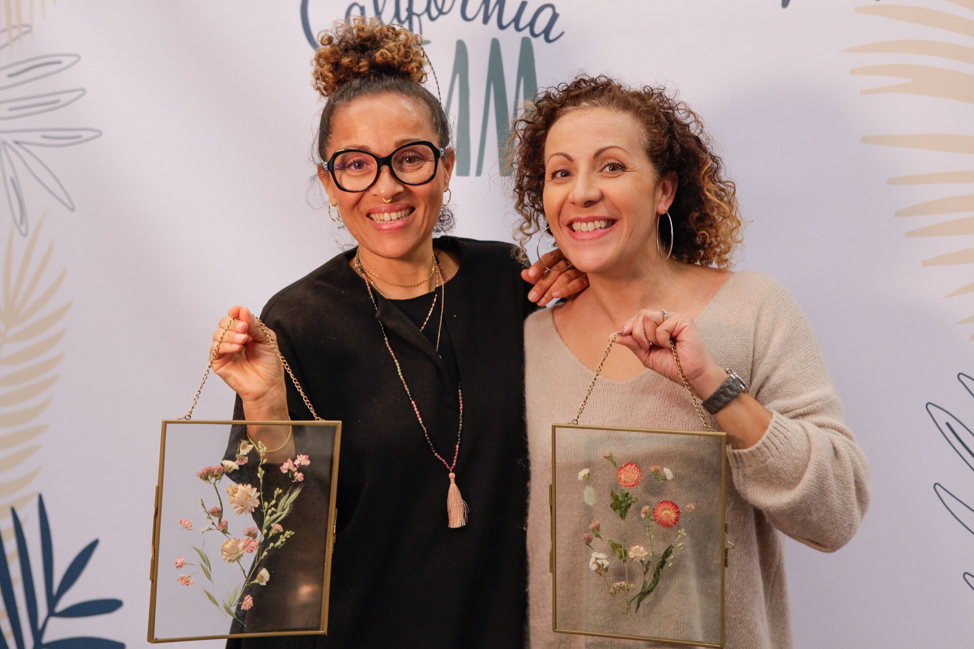 Deux femmes souriantes tenant des cadres avec des fleurs gravées. arrière-plan blanc avec motifs floraux et texte "California M".
