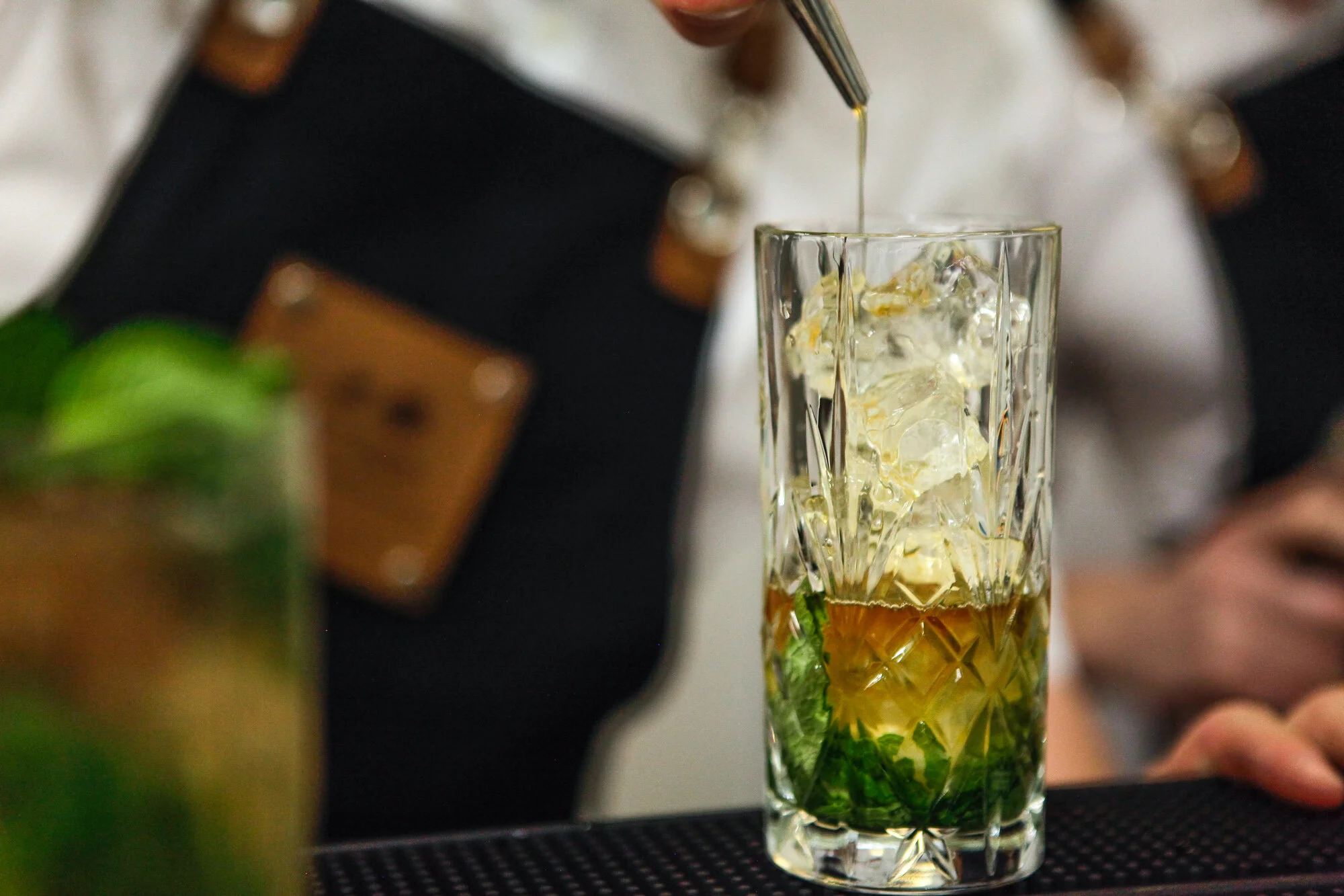 Une main verse de la liqueur dans un cocktail avec de la glace, de la menthe et du citron dans un verre en cristal.