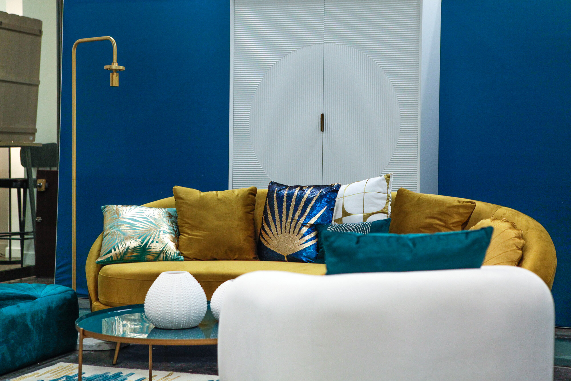 Un salon moderne avec un canapé jaune ornée de plusieurs coussins colorés et décoratifs, un tapis bleu, deux vases blancs sur une table ronde, un mur bleu en arrière-plan, et une porte blanche avec un motif circular.