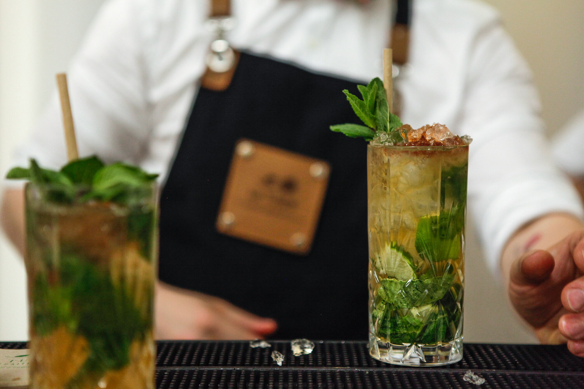 Un barman prépare un cocktail à base de menthe dans un verre haut avec de la glace, du citron, et une garniture de menthe, avec un autre verre en avant et un arrière-plan flou.