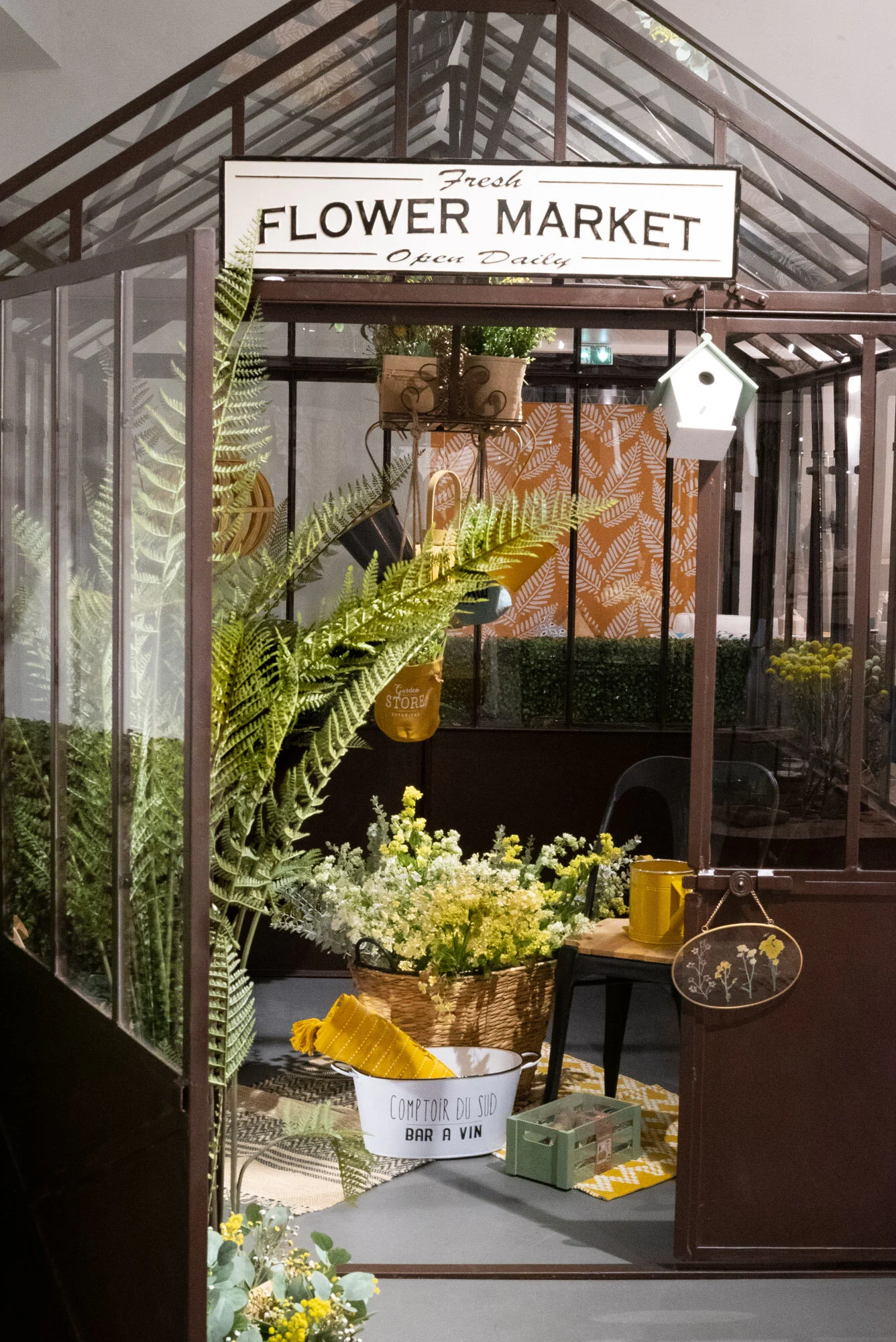 Une petite serre en verre avec une enseigne 'FRESH FLOWER MARKET' à l'entrée, remplie de plantes vertes, fleurs et objets de décoration.