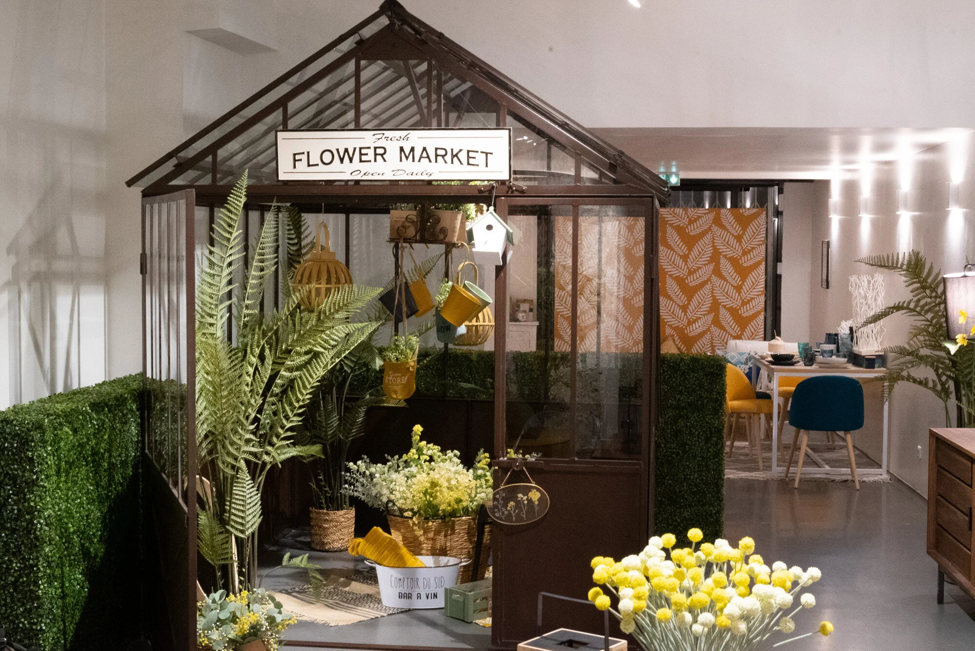 Petite serre en structure en métal noir avec panneau « Fleur Market » en haut, décoration florale avec plantes vertes et fleurs dans des paniers et pots, cuisine et salle à manger modernes en arrière-plan.
