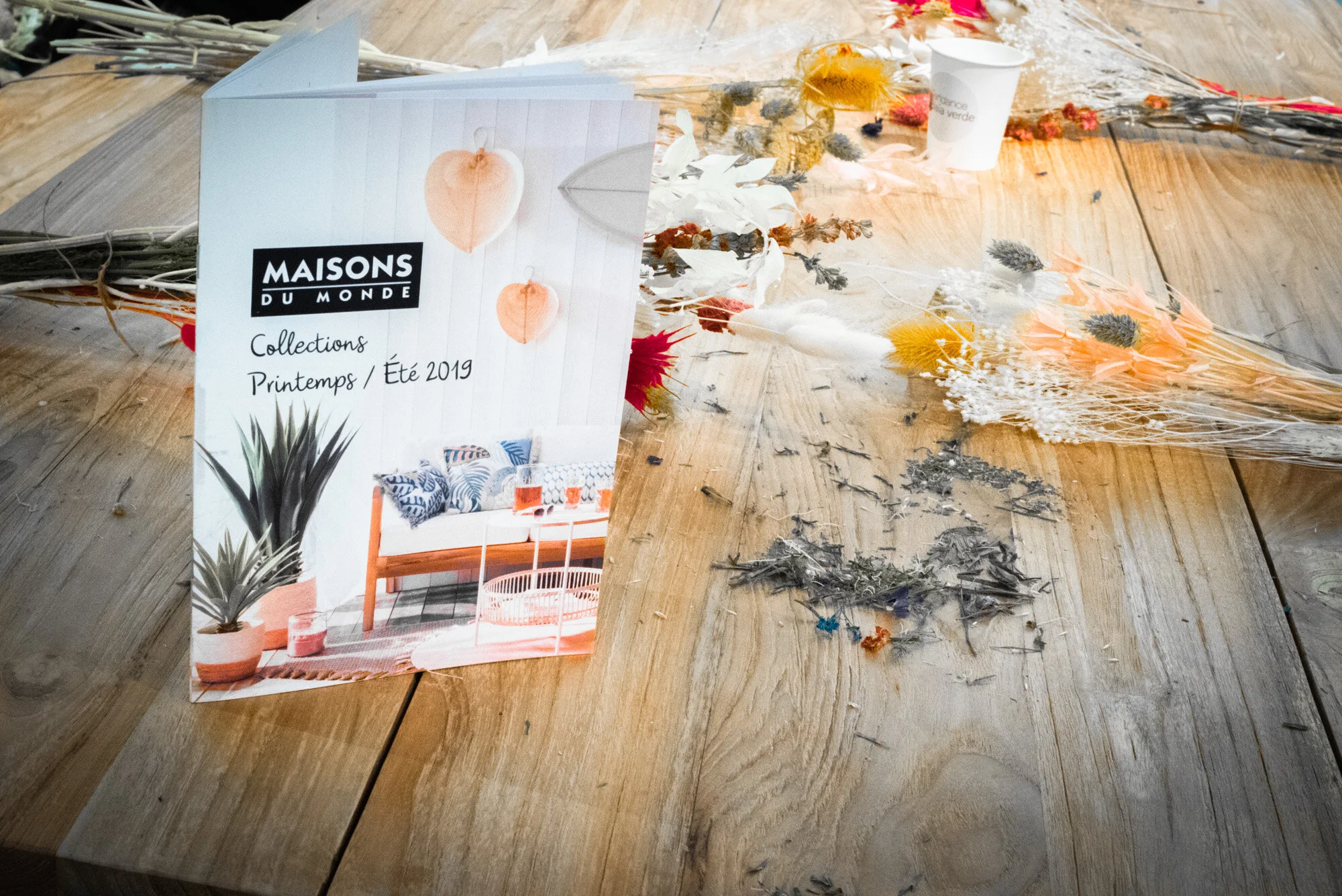 Une brochure promotionnelle de Maisons du Monde posée sur une table en bois, entourée de fleurs séchées