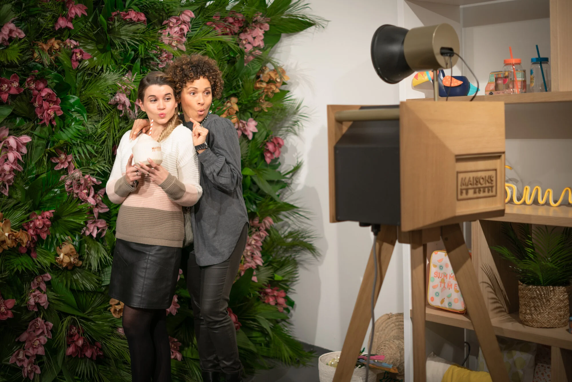 Deux femmes prennent une photo d'elles avec un fond de plantes et fleurs. Elles tiennent un verre avec une paille, l'une d'elles semble faire une expression surprise.