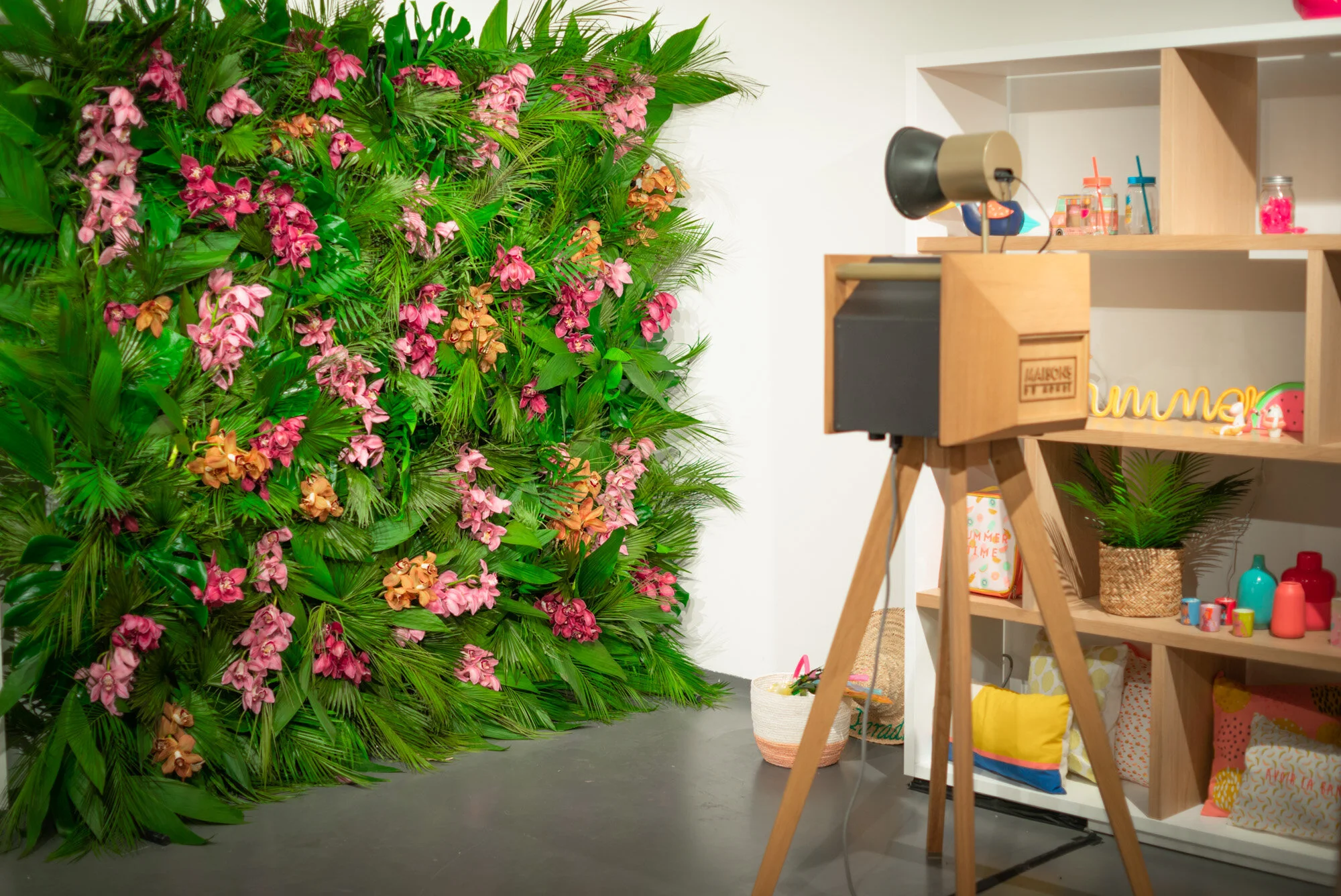 Un mur végétal avec des fleurs roses et des feuilles vertes. À droite, une étagère en bois avec divers objets décoratifs, y compris un projecteur, des petites figurines et des bocaux colorés.