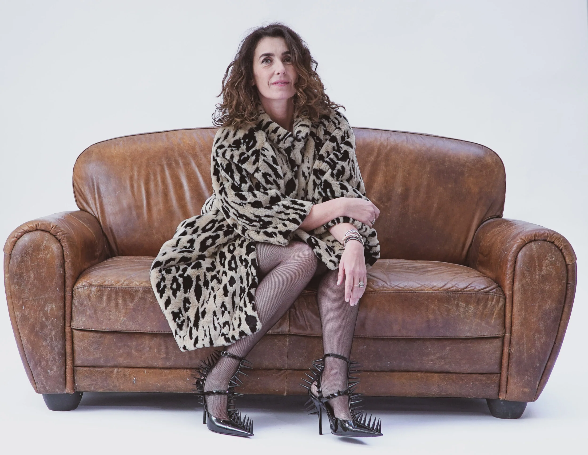 Femme assise sur un canapé en cuir brun, portant un manteau à imprimé léopard, des collants gris et des chaussures à talons noirs avec des pointes et des fermetures à boucles.