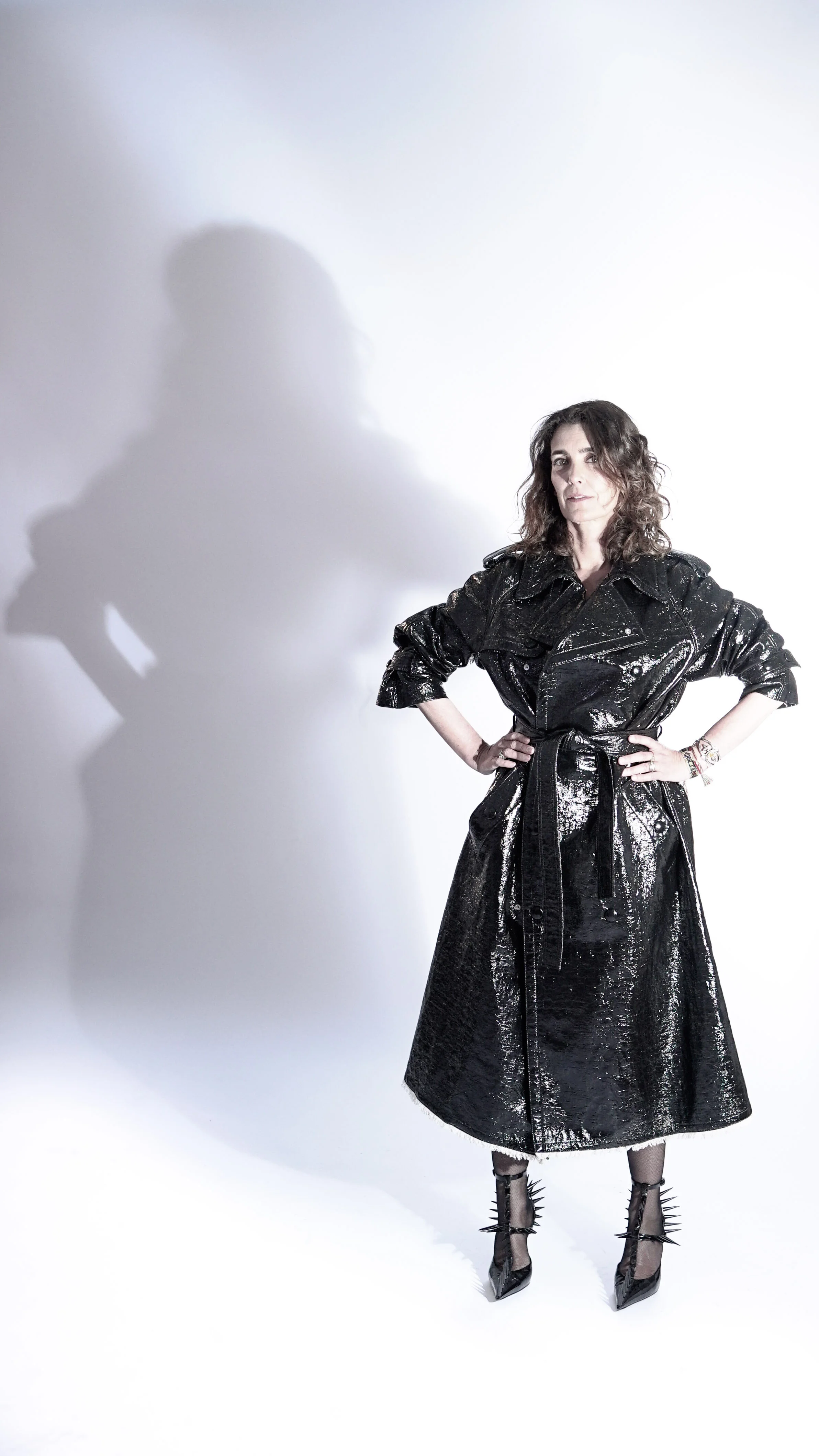 Une femme posant avec une main sur la hanche, portant un manteau noir brillant et des chaussures à talons aiguilles ornées de piquants, sous un éclairage dramatique créant une ombre sur le mur blanc derrière elle.