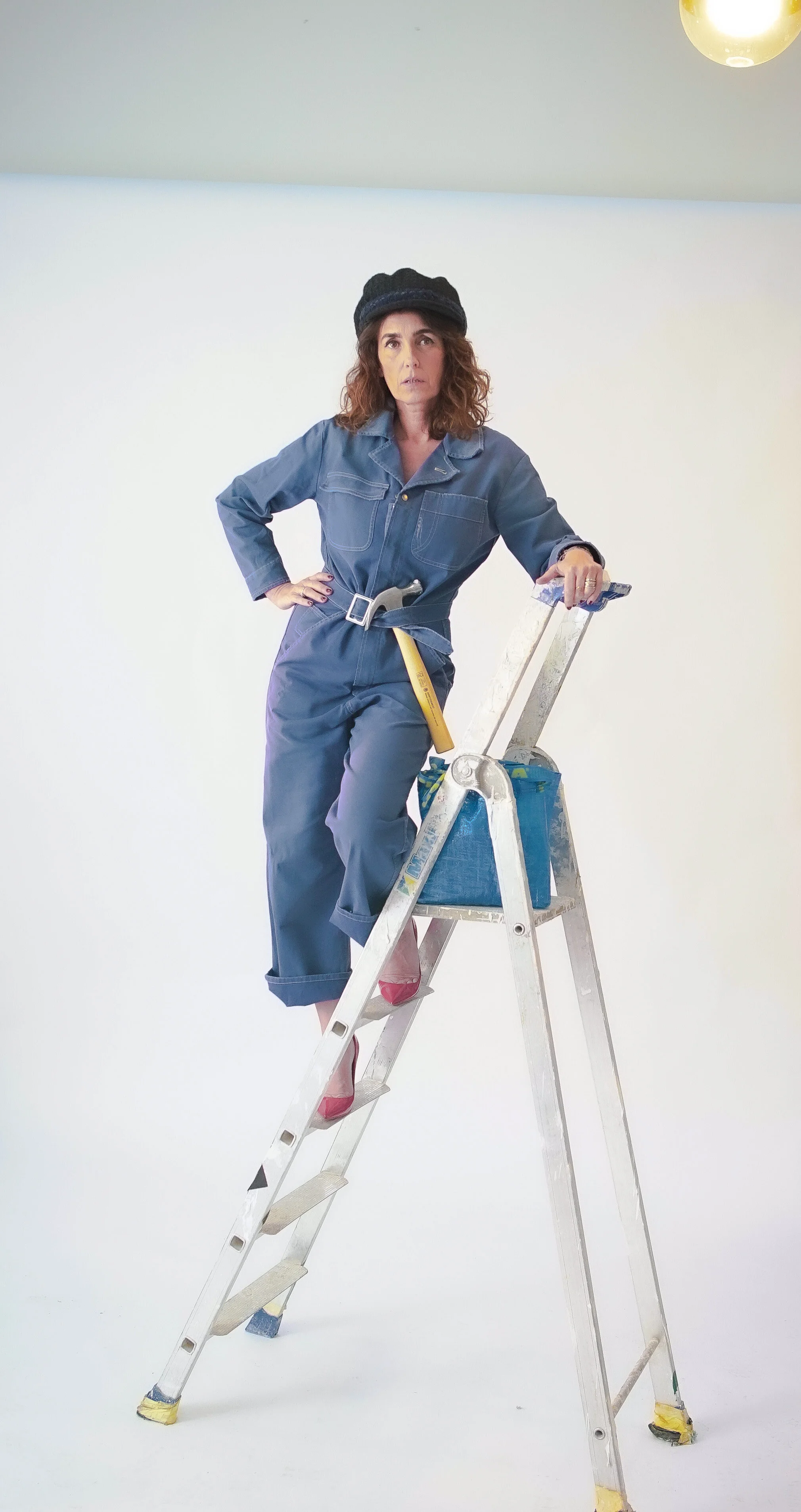 Femme posant sur une échelle, portant une combinaison de travail bleue et un béret noir, avec une panière à outils.