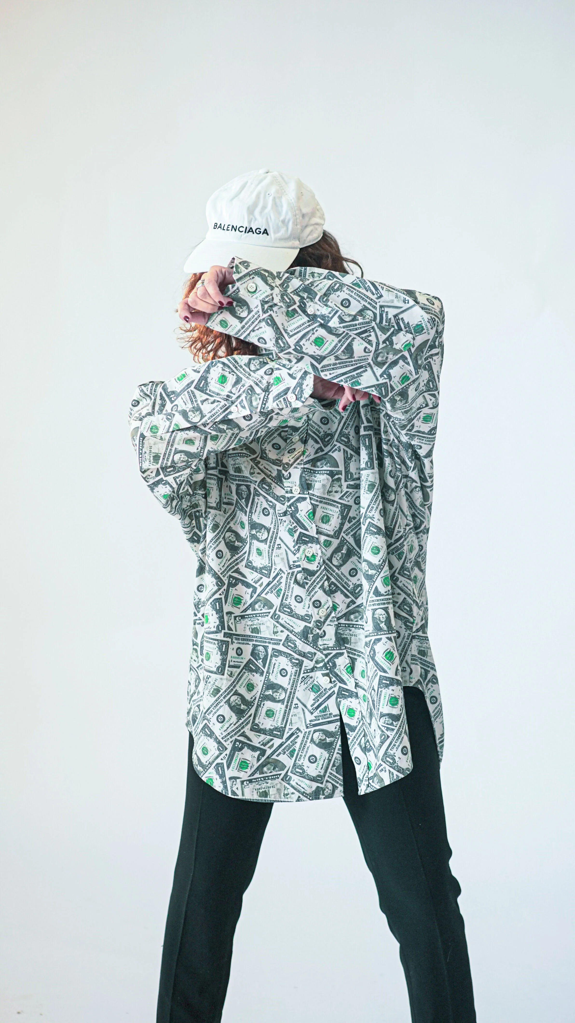 Une personne portant une chemise et un chapeau, tous deux couverts de motifs de dollars, se couvre partiellement le visage avec la main.