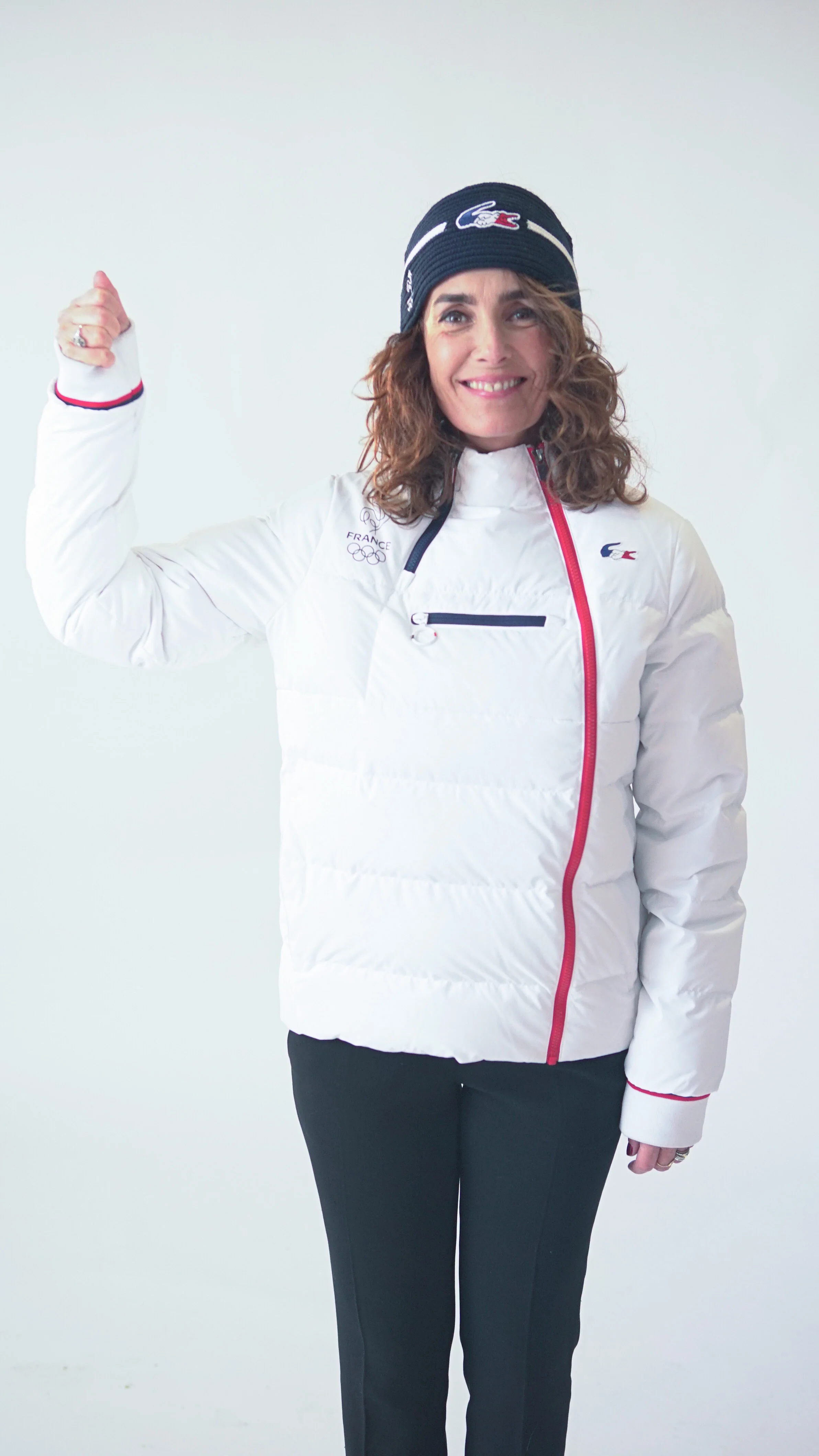 Une femme souriante en tenue de sport blanche avec un bonnet noir, faisant un signe de victoire, contre un fond blanc.