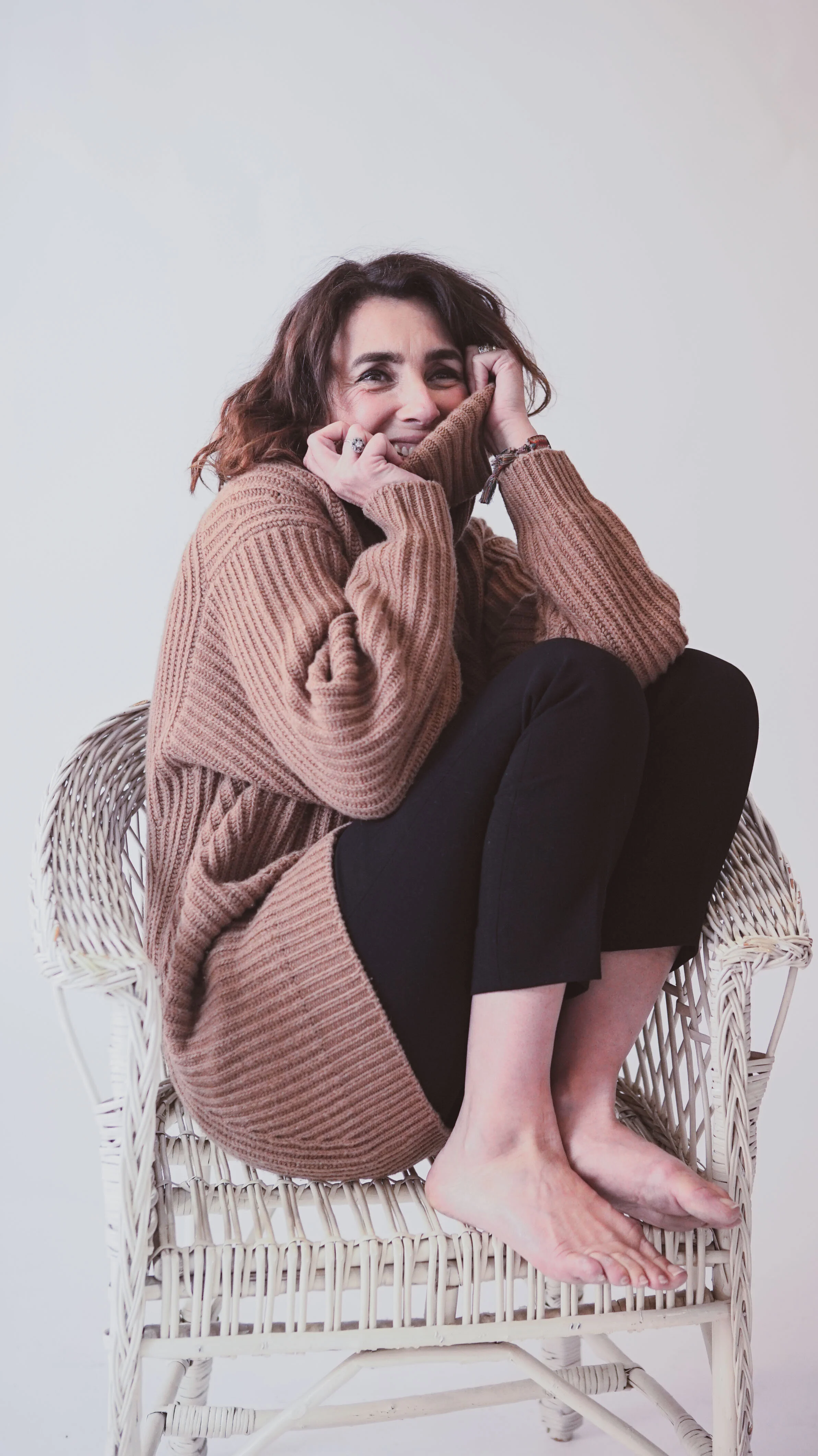 Une femme assise dans un fauteuil en osier, portant un pull marron, souriant et se cachant partiellement le visage avec ses mains.