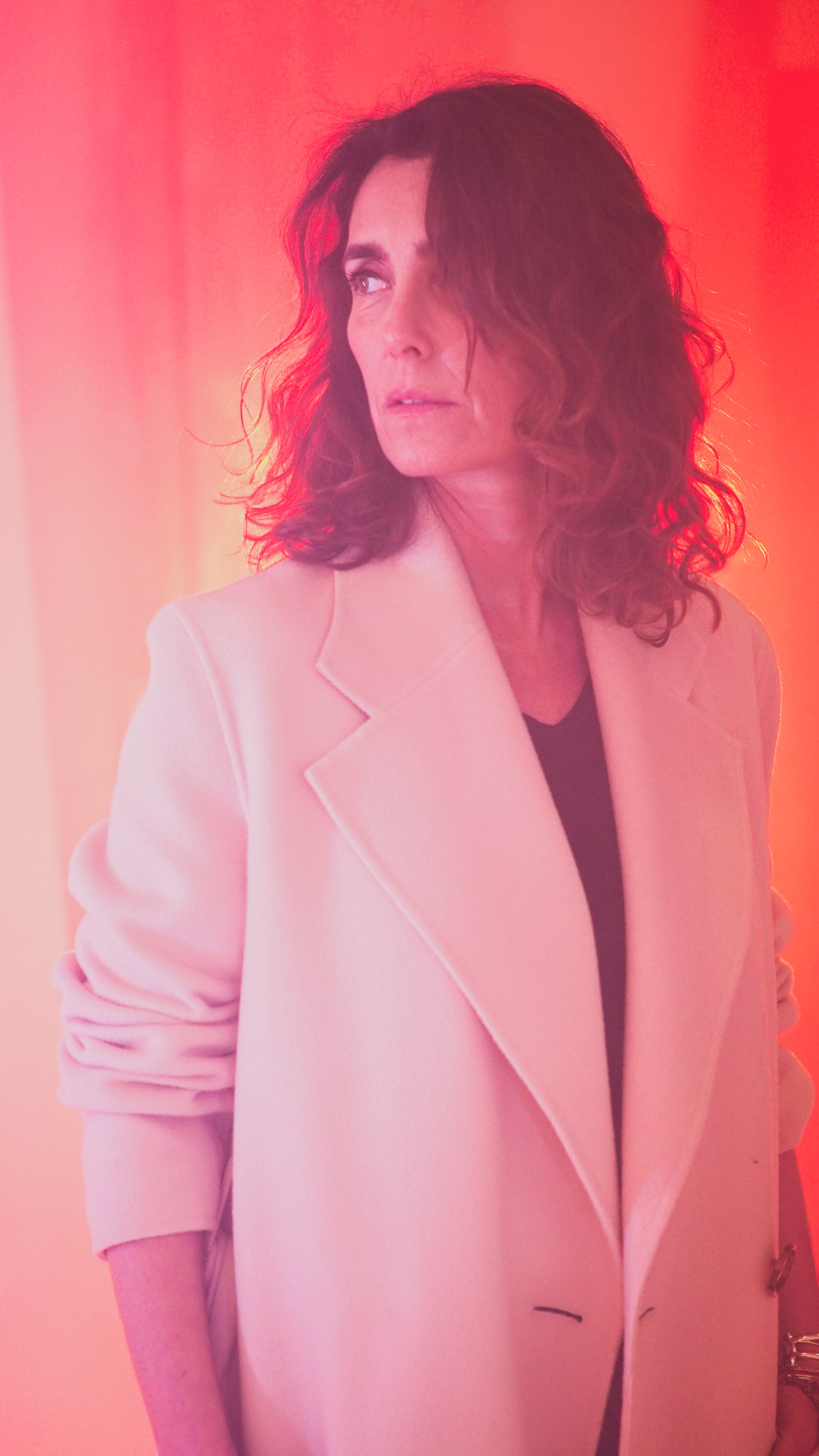 Une femme aux cheveux bouclés, portant un blazer rose pâle, regarde vers la gauche, avec un fond rouge-orange.