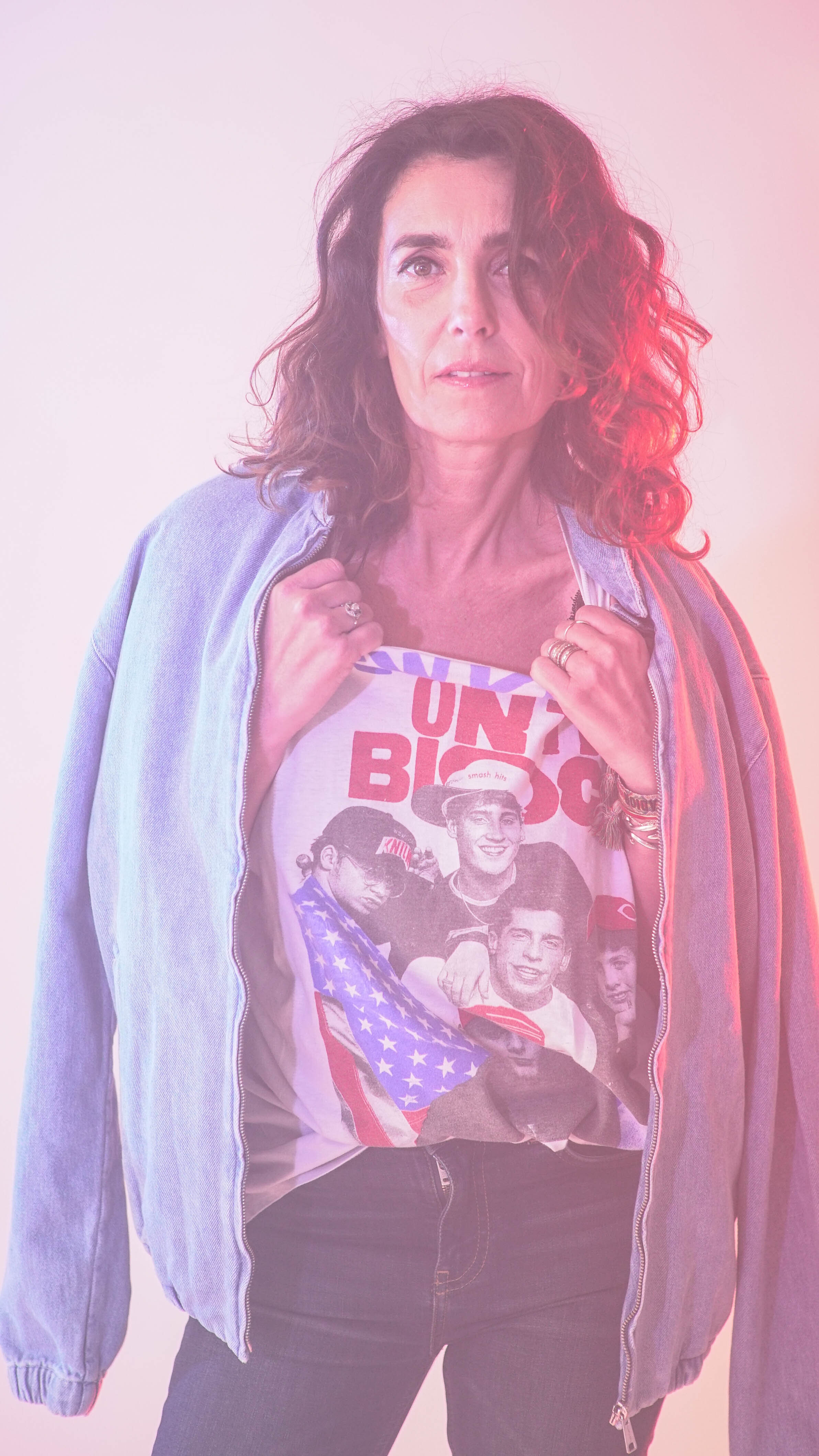 Femme portant un t-shirt avec une image de plusieurs jeunes gens et un drapeau américain, en pull bleu à capuche. Elle a les cheveux bouclés et porte des bracelets.