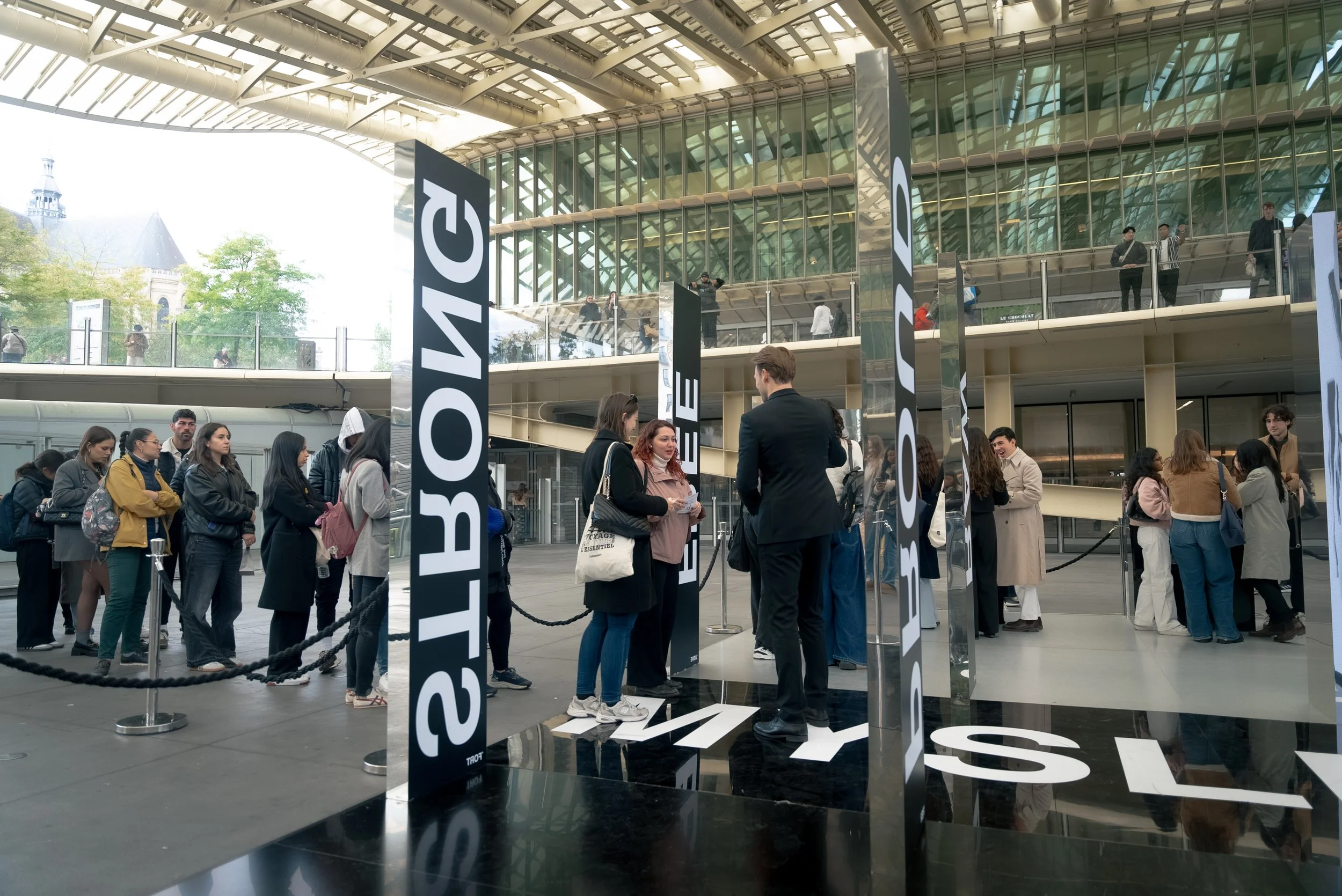 Une file d'attente devant une entrée d'un bâtiment moderne avec des panneaux indiquant 'STUDIO' et 'RÉSERVER'. Des personnes attendent ou communiquent, avec certains couverts par une barrière. La structure en verre et métal du bâtiment est visible en