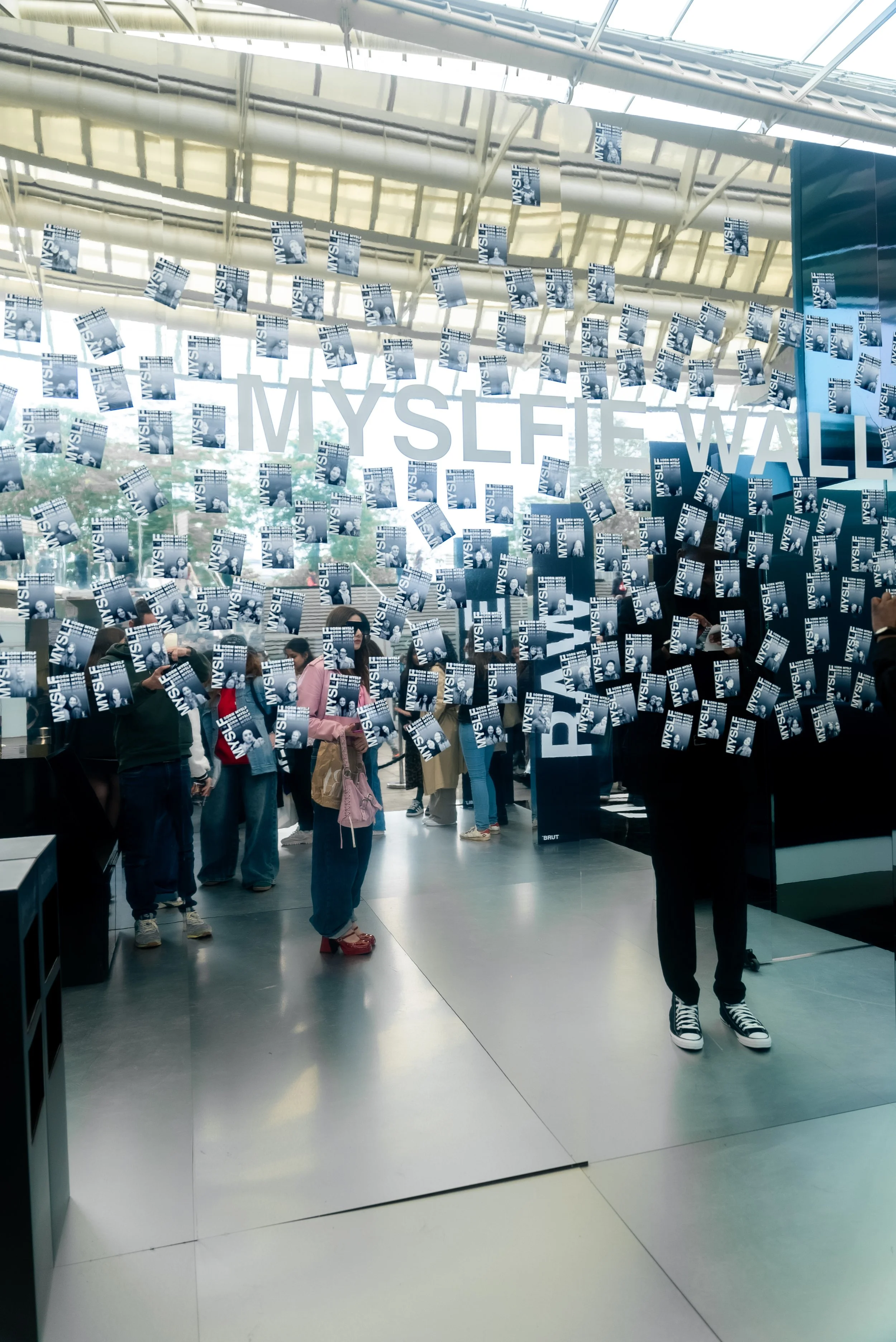 Exposition d'art interactif avec des photos suspendues, la phrase 'MYSSELF' visible en arrière-plan, des visiteurs dans un espace moderne avec une grande verrière.