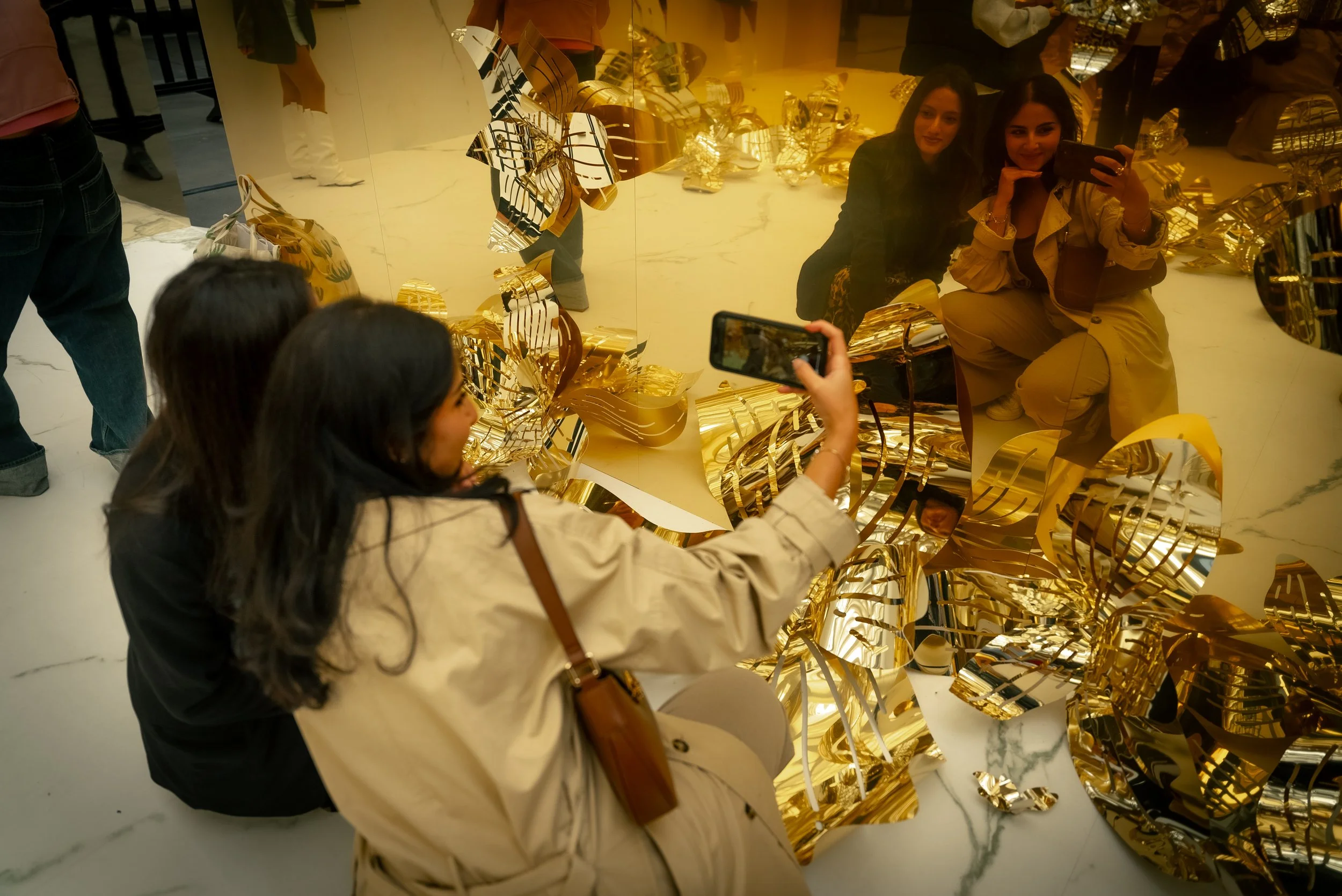 Deux femmes posent devant un miroir doré dans une boutique, tandis qu'une autre prend une photo d'elles avec son téléphone. L'ensemble est entouré de décorations en feuilles dorées.