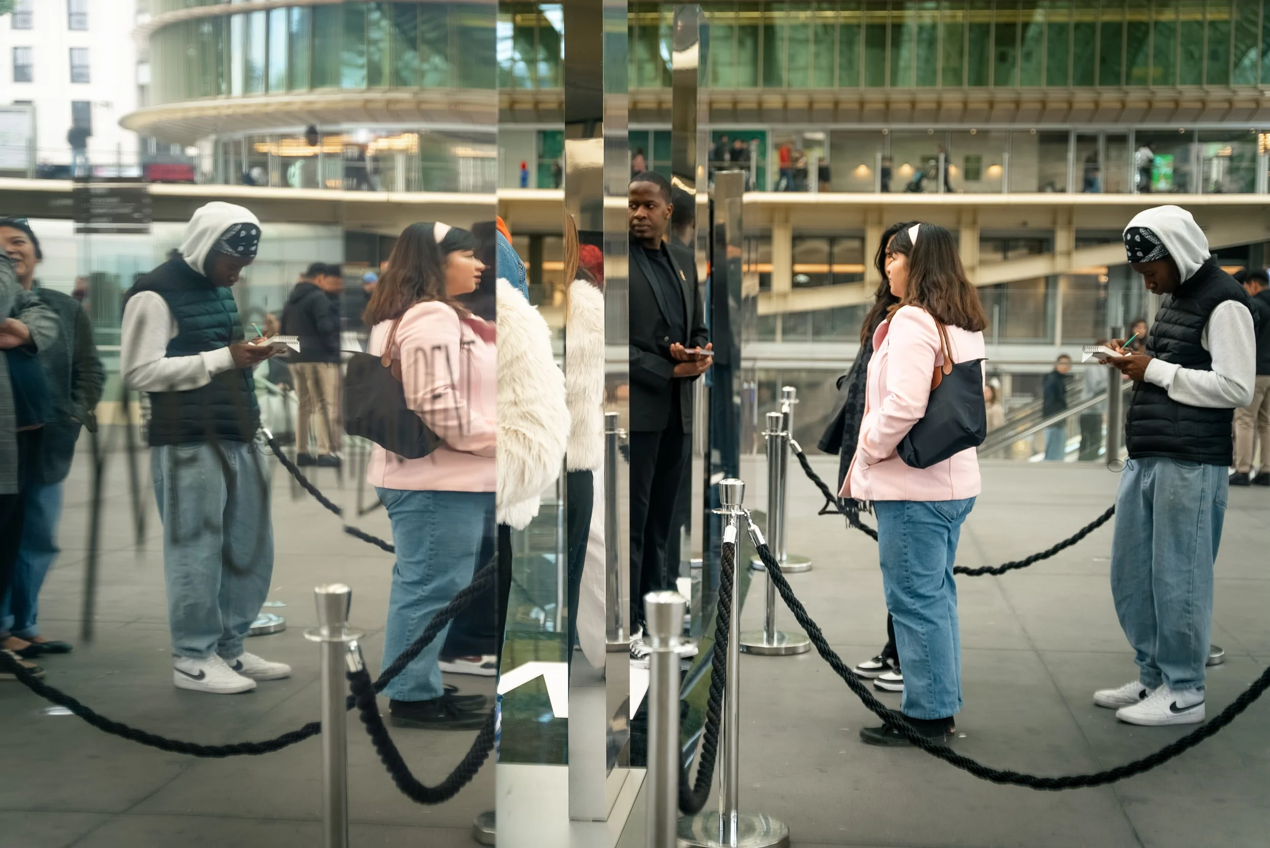 Reflet d'une file d'attente de personnes devant un bâtiment moderne en verre, avec plusieurs jeunes utilisant leur téléphone, certains faisant la queue et d'autres regardant leur téléphone.