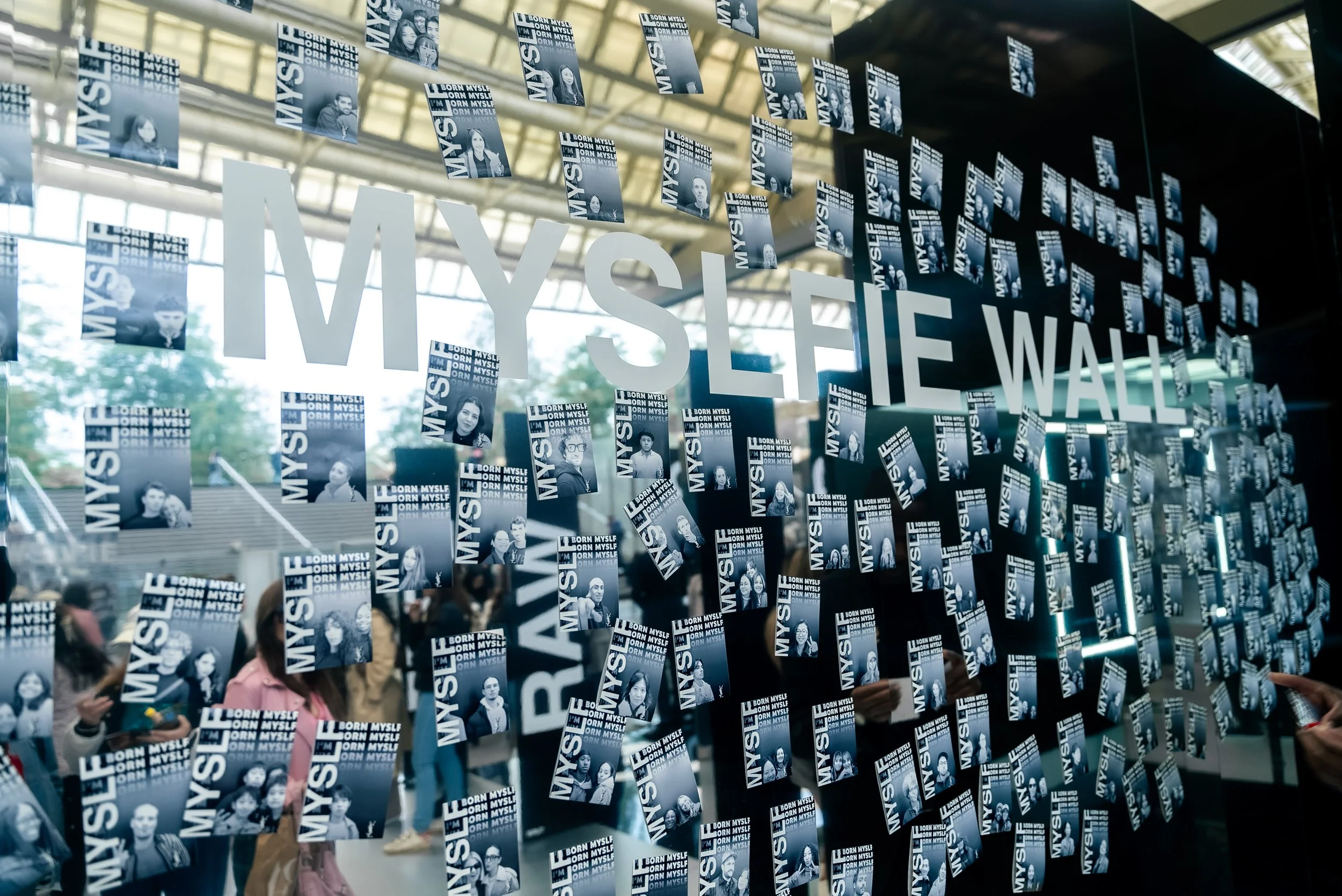 Vitrine d'exposition avec le texte 'I MYSELF WALL' et de nombreuses petites affiches avec des portraits de personnes et la phrase 'BORN MYSELF'.