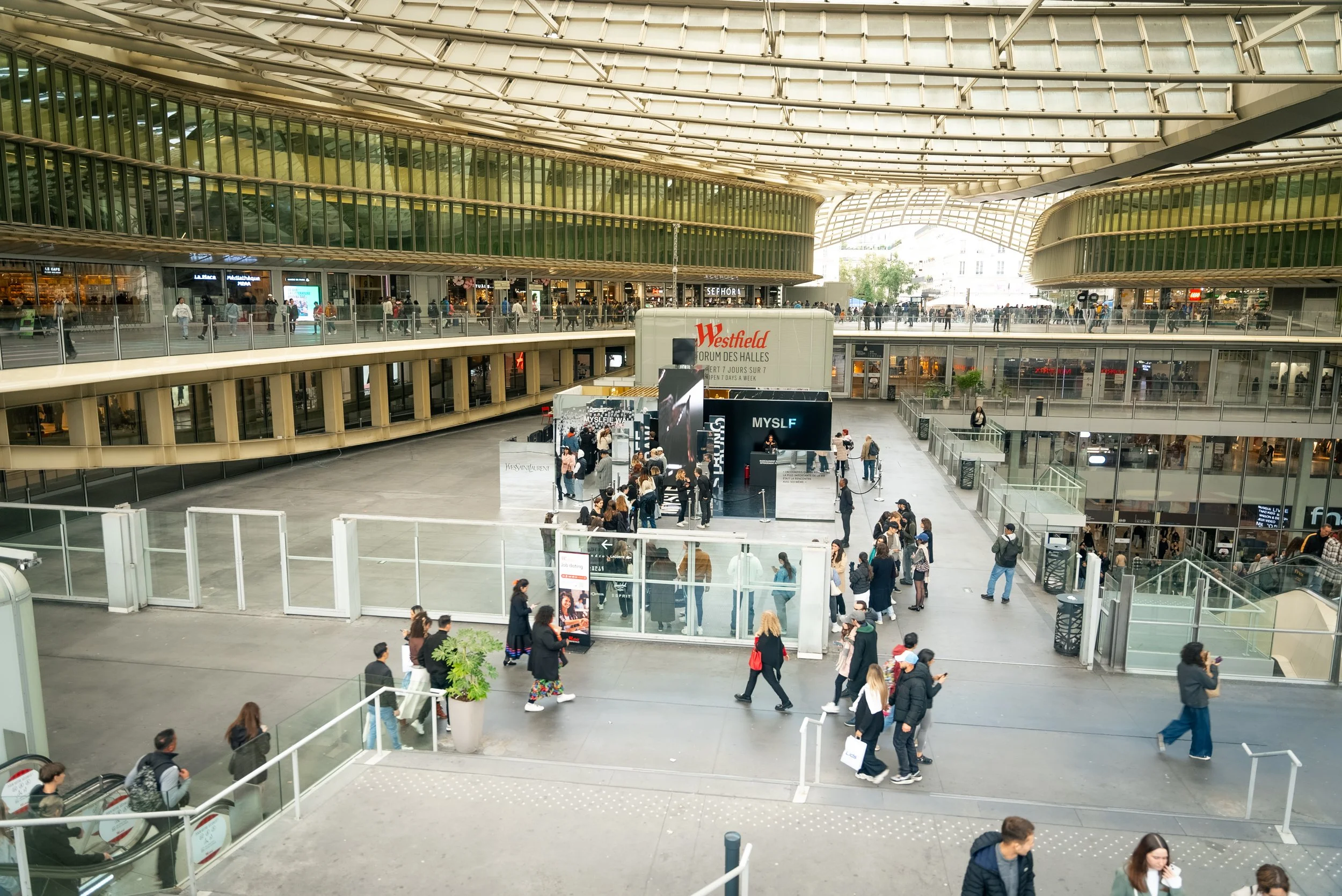 Vue intérieure d'un centre commercial avec plusieurs niveaux, escalators, et des personnes marchant ou utilisant leurs téléphones, sous une grande structure en verre.