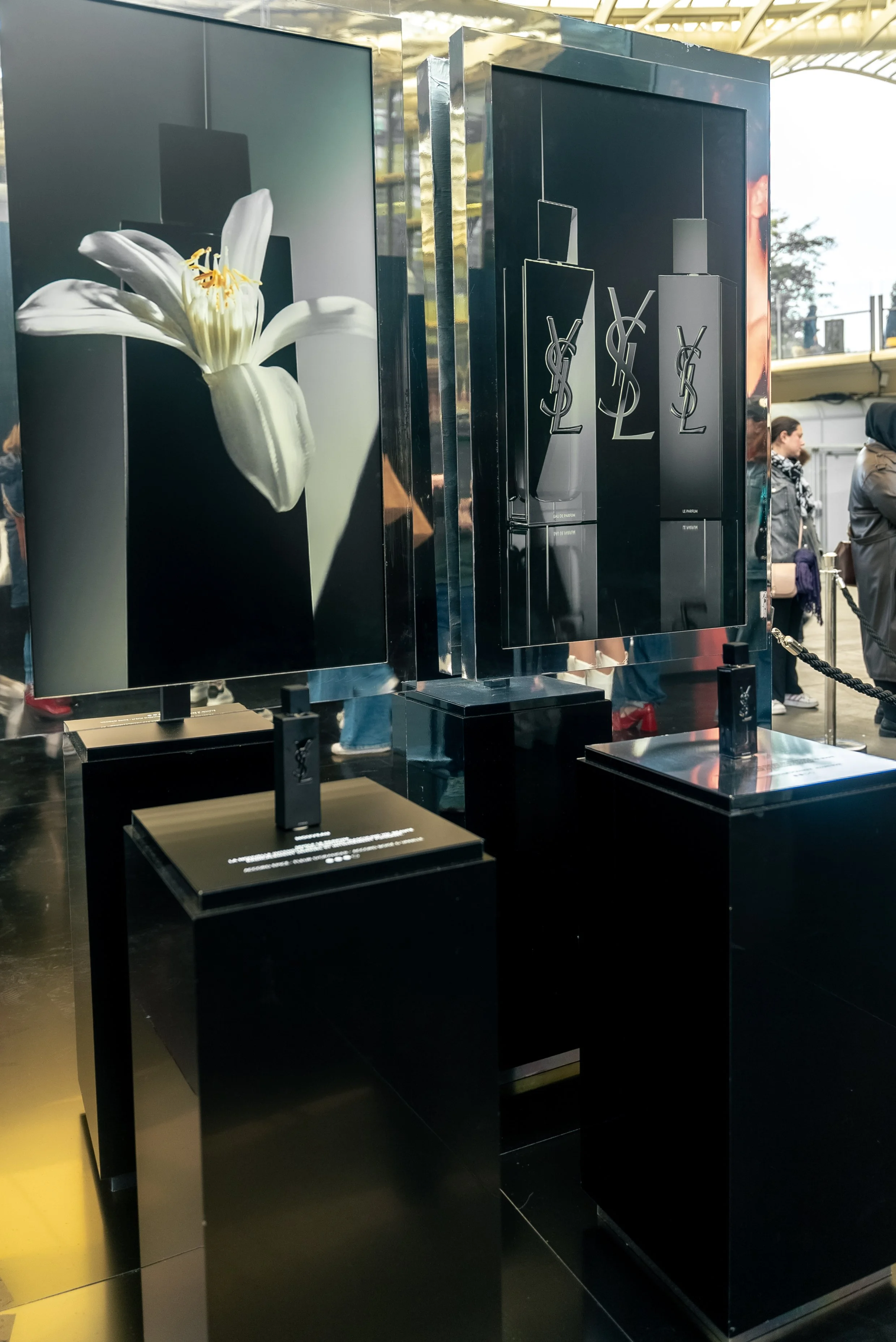 Vitrine de publicité pour la marque Yves Saint Laurent, avec une image d'une fleur blanche et des bouteilles de parfum noires au design minimaliste.