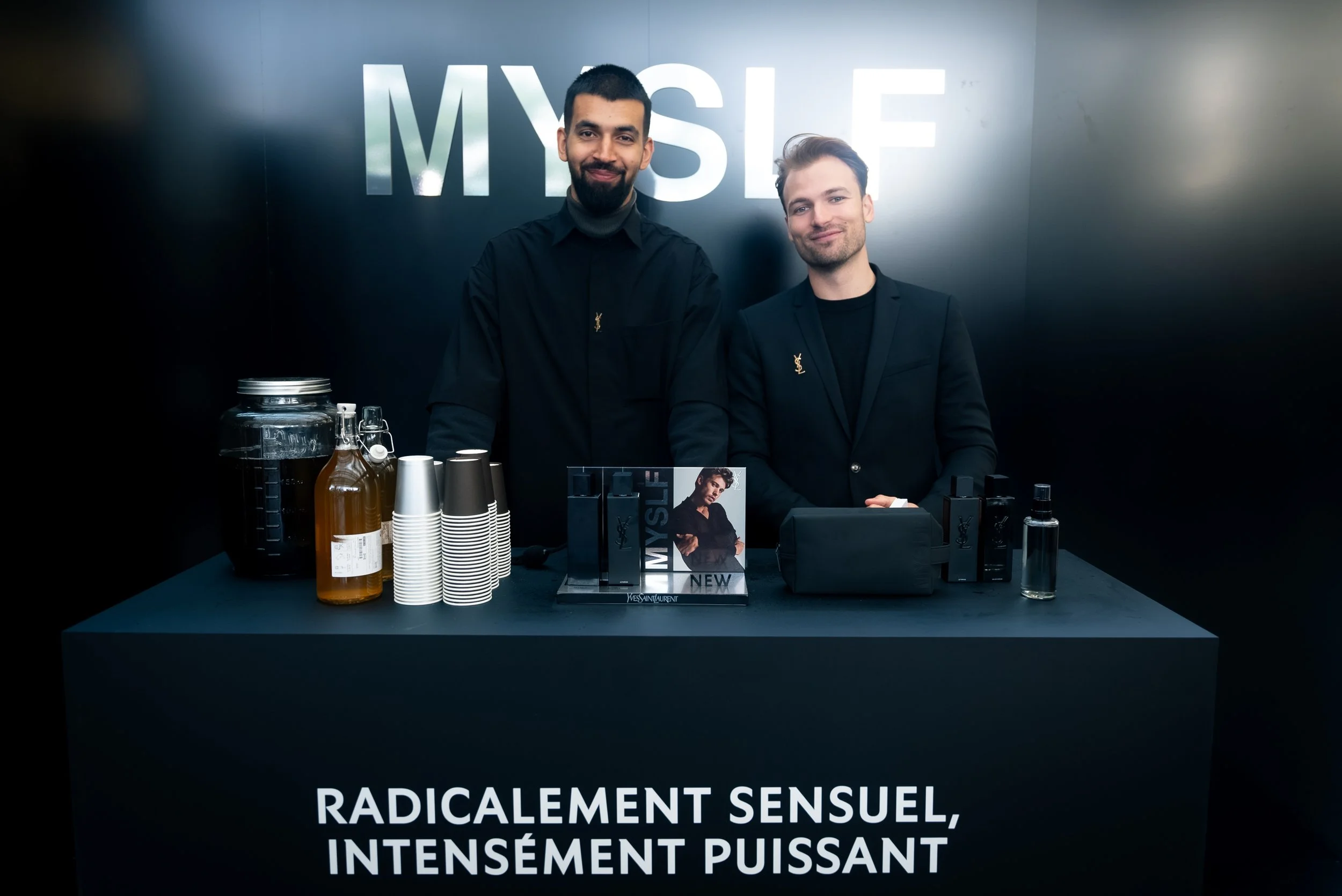 Deux hommes en tenue noire se tiennent derrière un comptoir de présentation de produits Yves Saint Laurent, avec des bouteilles, des gobelets en papier, un cadre avec une affiche, et des flacons de parfum, sur un fond noir avec le mot 'MYSLE' en gros