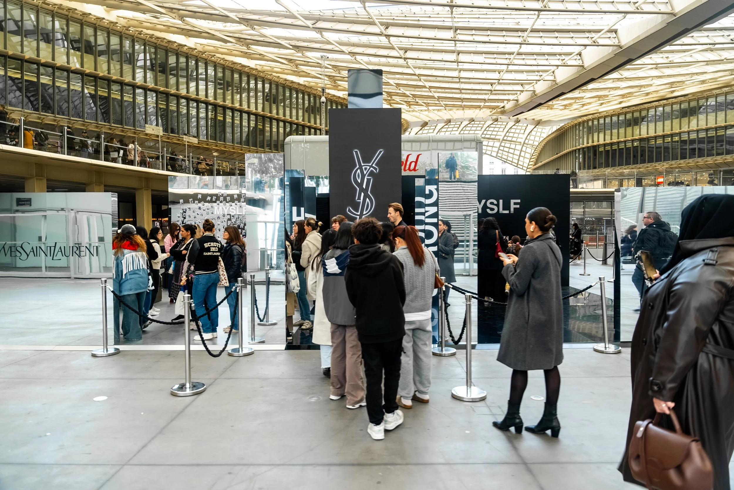 Groupe de personnes faisant la queue devant une attraction de marque Yves Saint Laurent dans un centre commercial moderne.