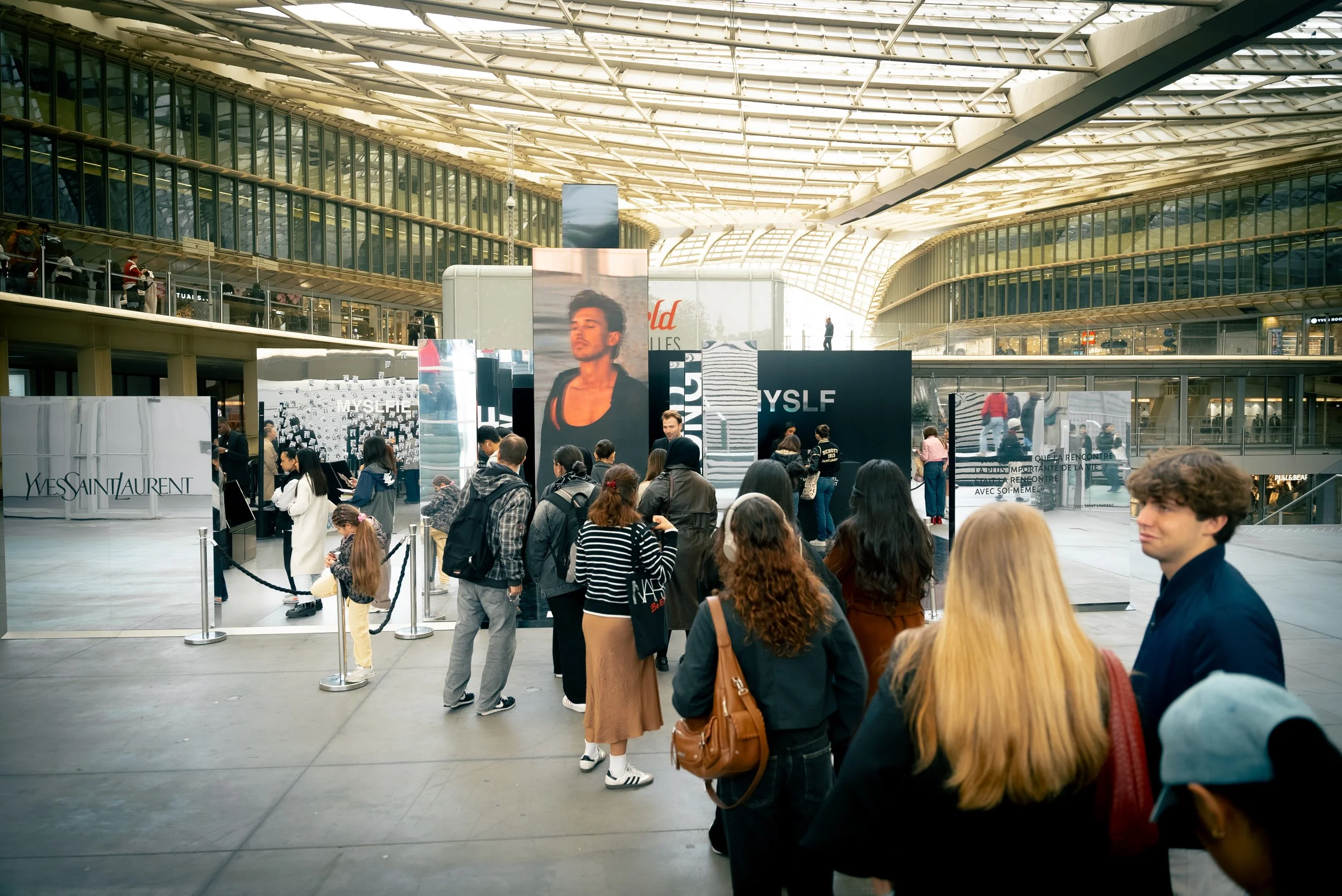 Une foule de personnes attendant dans une galerie commerciale moderne avec un plafond en verre, à côté des stands de marques telles que Yves Saint Laurent.