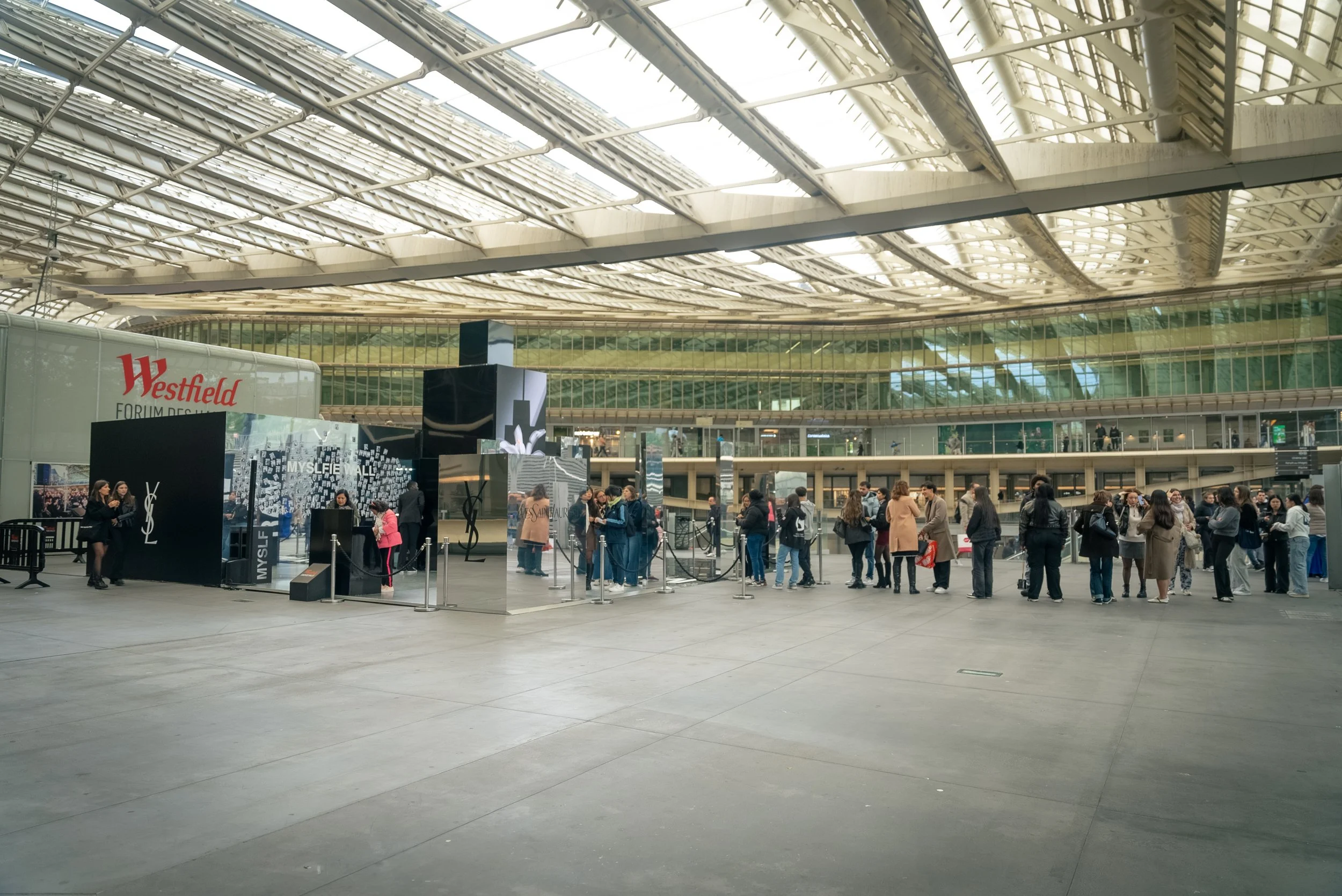 Une file d'attente de personnes devant un stand de Laurent en intérieur dans un centre commercial ou une exposition avec un plafond en verre et une structure métallique.