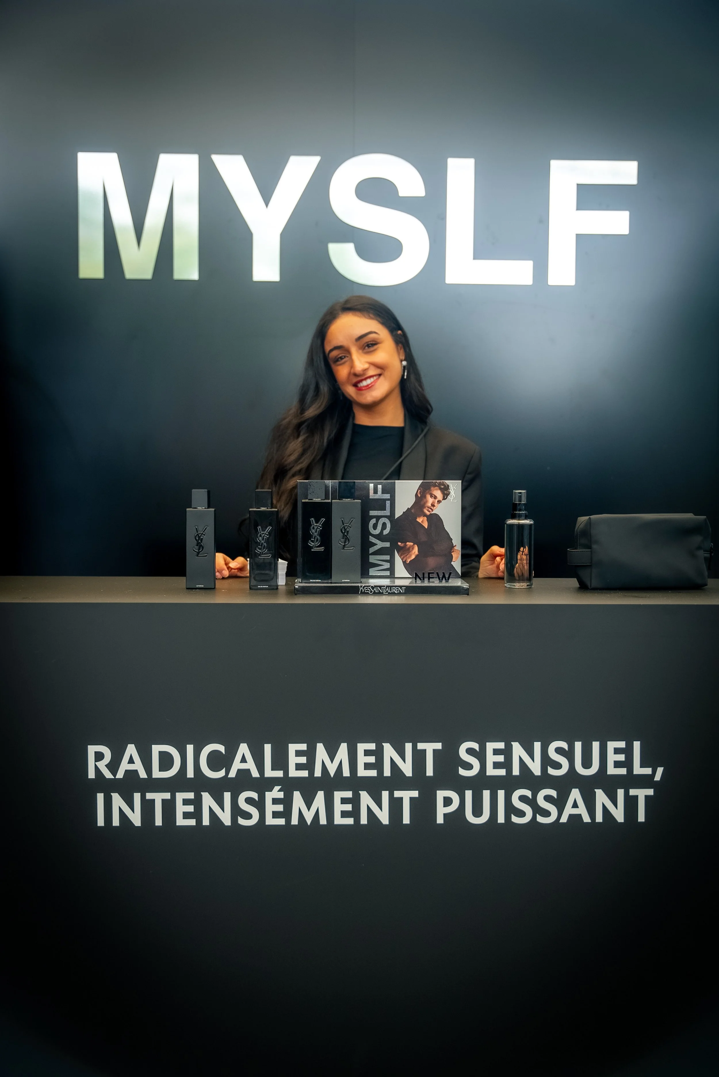 Une femme souriante se tient derrière un stand de promotion avec le mot "MYSLE" en lettres lumineuses derrière elle. Sur le stand, il y a des flacons de parfum, un coffret, et un sac noir. Le panneau en dessous indique : "RADICALEMENT SENSUEL, INTENS