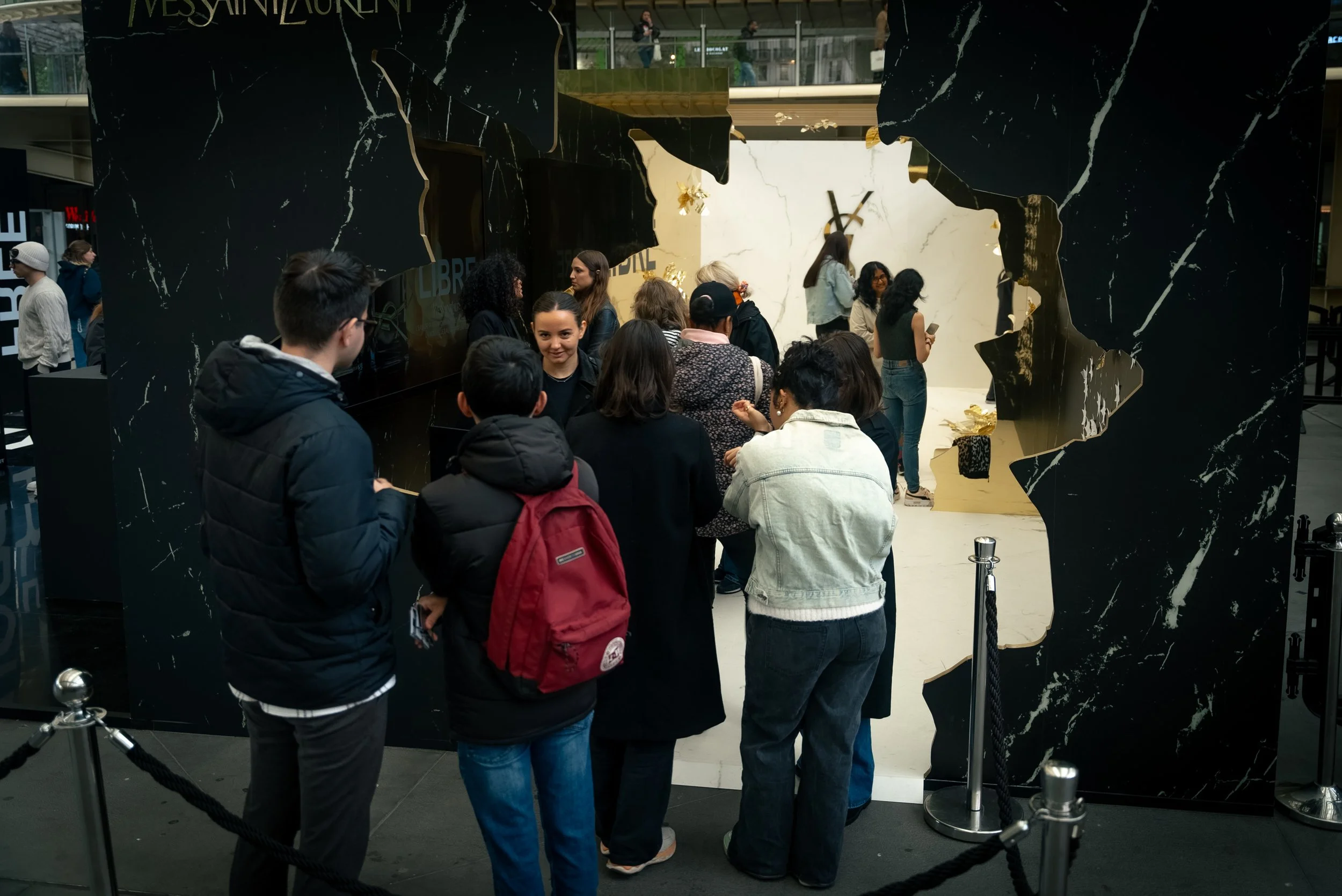 Groupe de personnes attendant à une file d'attente devant un comptoir à l'intérieur d'un centre commercial ou une boutique, avec un mur noir marbré et un espace en forme de trou derrière eux.